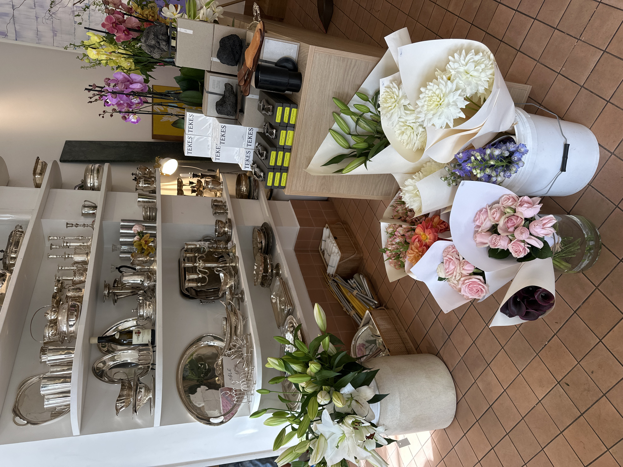 The image captures a storefront in Tel-Aviv, Israel. Inside, a well-lit shop features an assortment of merchandise. On the left, white shelves display polished silverware, including trays, teapots, and other serving pieces. The middle section showcases floral arrangements, including orchids and lilies. A white bucket holds a bouquet of white dahlias and blue flowers. Other bouquets of pink roses and dark purple calla lilies rest on the tiled floor. Boxes labeled "TEKES TEKES" are stacked on a shelf.