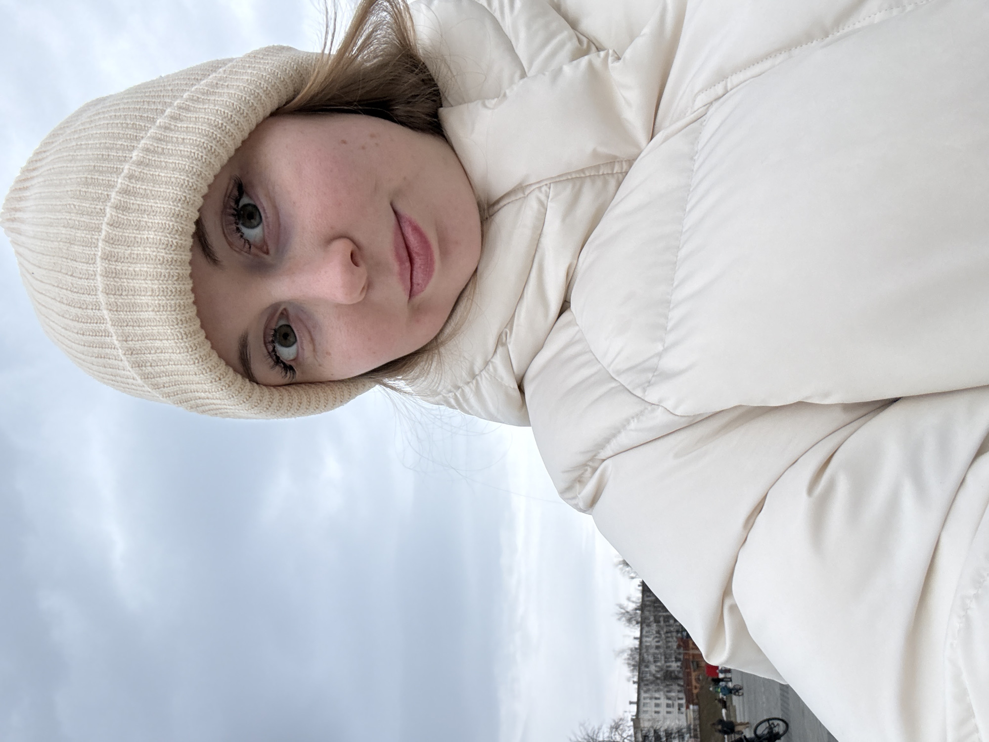 A fair-skinned young woman with light-colored hair and green eyes is seen in a selfie. She is wearing a light-colored beanie and a puffy white jacket. The background features a cloudy sky, and in the distance, there is a building, some trees, and a few people riding bicycles.