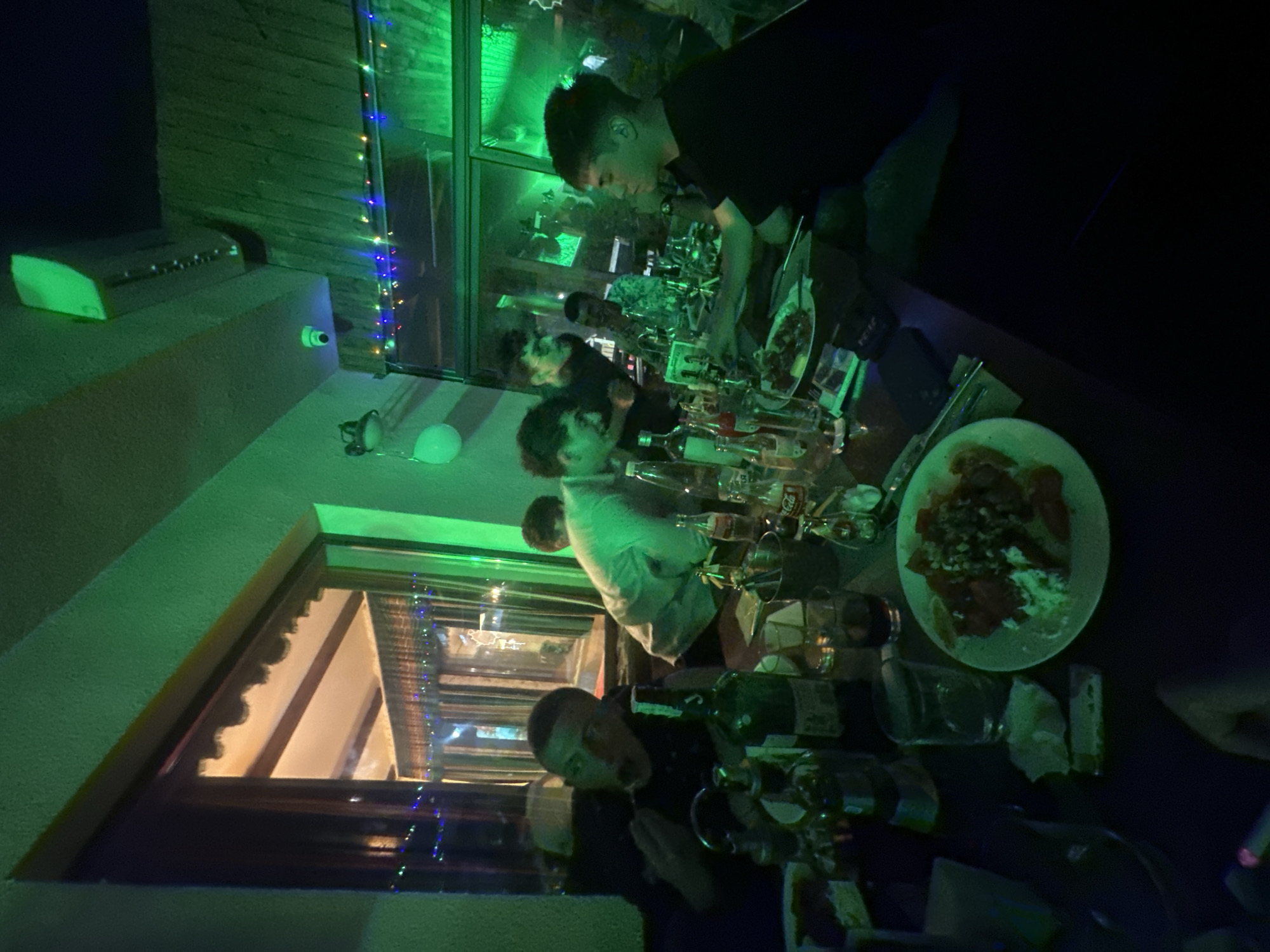 A medium indoor shot shows a crowded table setting, presumably in a restaurant or bar. Several people are seated around the table, seemingly engaged in a meal or social gathering. The lighting has a strong green hue, casting a distinctive tone over the scene. A plate of food, possibly a salad, is prominently placed on the table. Numerous empty bottles and glassware are scattered around, indicating consumption. A window with decorations and a partial view of an illuminated exterior suggests that this location might be in the city of Panagyurski kolonii, Bulgaria.