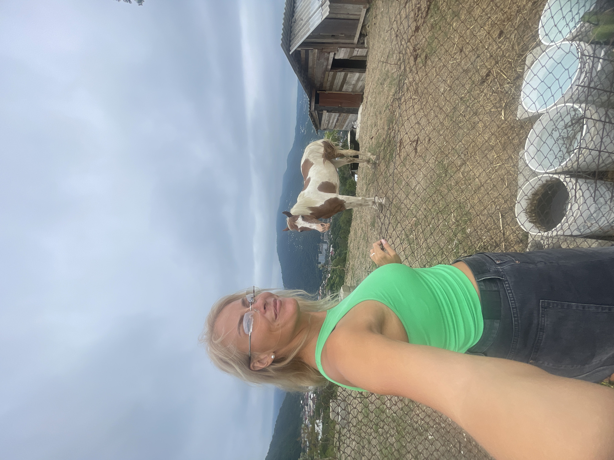 In Sochi, Russia, a blonde woman with glasses and a green tank top is taking a selfie in front of a brown and white horse. The horse is standing next to a rustic wooden structure with a corrugated metal roof. The background shows a hilly, green landscape with buildings visible in the distance, under a cloudy sky. The woman is positioned in the foreground on the left side of the frame, and the horse is in the middle ground on the right. A wire fence runs across the bottom of the image, separating the viewer from the scene. There are several white buckets visible behind the fence in the lower right corner. The overall environment suggests a rural or semi-rural setting within Sochi.