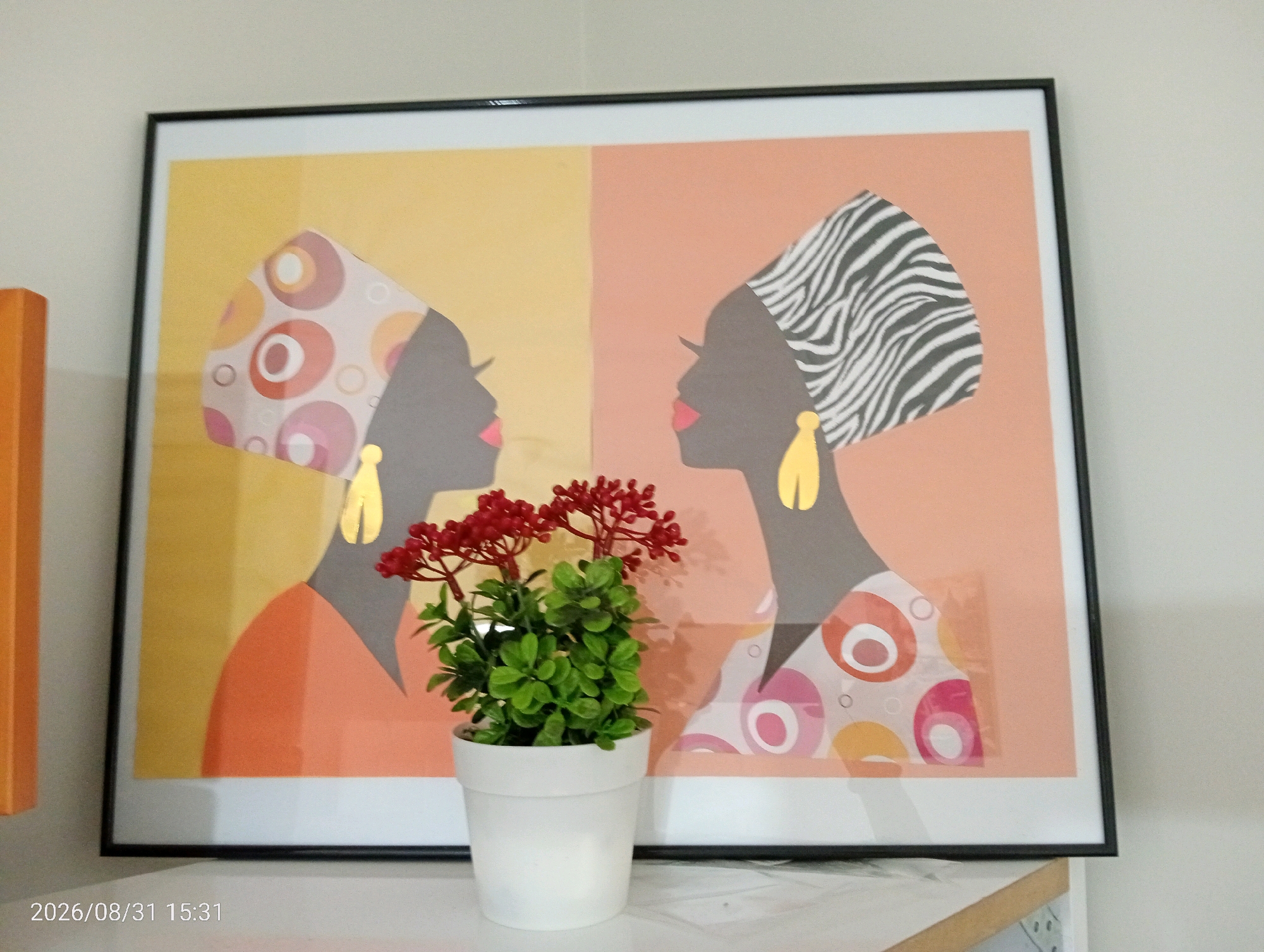 A framed artwork featuring two silhouetted profiles of women is displayed against a wall. The artwork is divided into two vertical sections of color: yellow on the left and peach on the right. Both women are wearing headwraps; the woman on the left has a headwrap with colorful circular patterns, and the woman on the right has a headwrap with a zebra-like pattern. Both wear large, yellow, teardrop-shaped earrings and have pink lips. In the foreground, in front of the artwork, is a small white pot containing a faux plant with green leaves and red berries. The bottom left corner of the image shows a timestamp: "2026/08/31 15:31". The scene appears to be indoors, on a shelf or table. There is no indication of the city of Silifke, Turkey in the image itself.