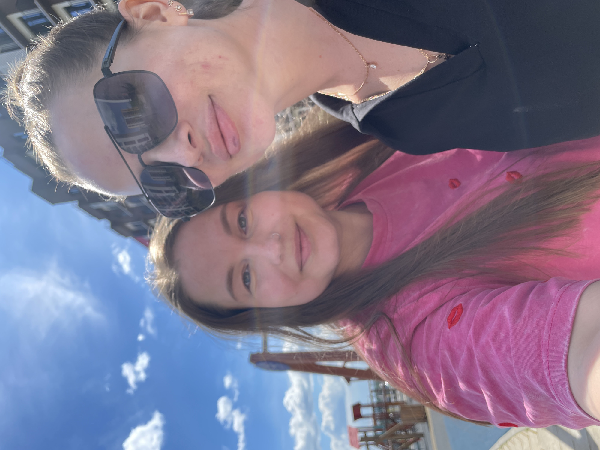 Two women are depicted in a close-up outdoor selfie under a bright blue sky with scattered white clouds. The woman on the left, with long brown hair, wears a pink t-shirt with red lip embroideries and a small nose piercing, smiling gently towards the camera. The woman on the right, partially visible, wears large dark sunglasses that reflect the sky and a distant building, a black collared top, and gold jewelry including a necklace with a clear stone pendant and small earrings. Her facial skin exhibits some red blemishes. In the blurred background, a children's playground with wooden structures and a red roof is visible to the left, and a modern multi-story building with dark windows stands to the right. No specific identifying markers for the city of Pervomaysky, Russia, are present in the image.