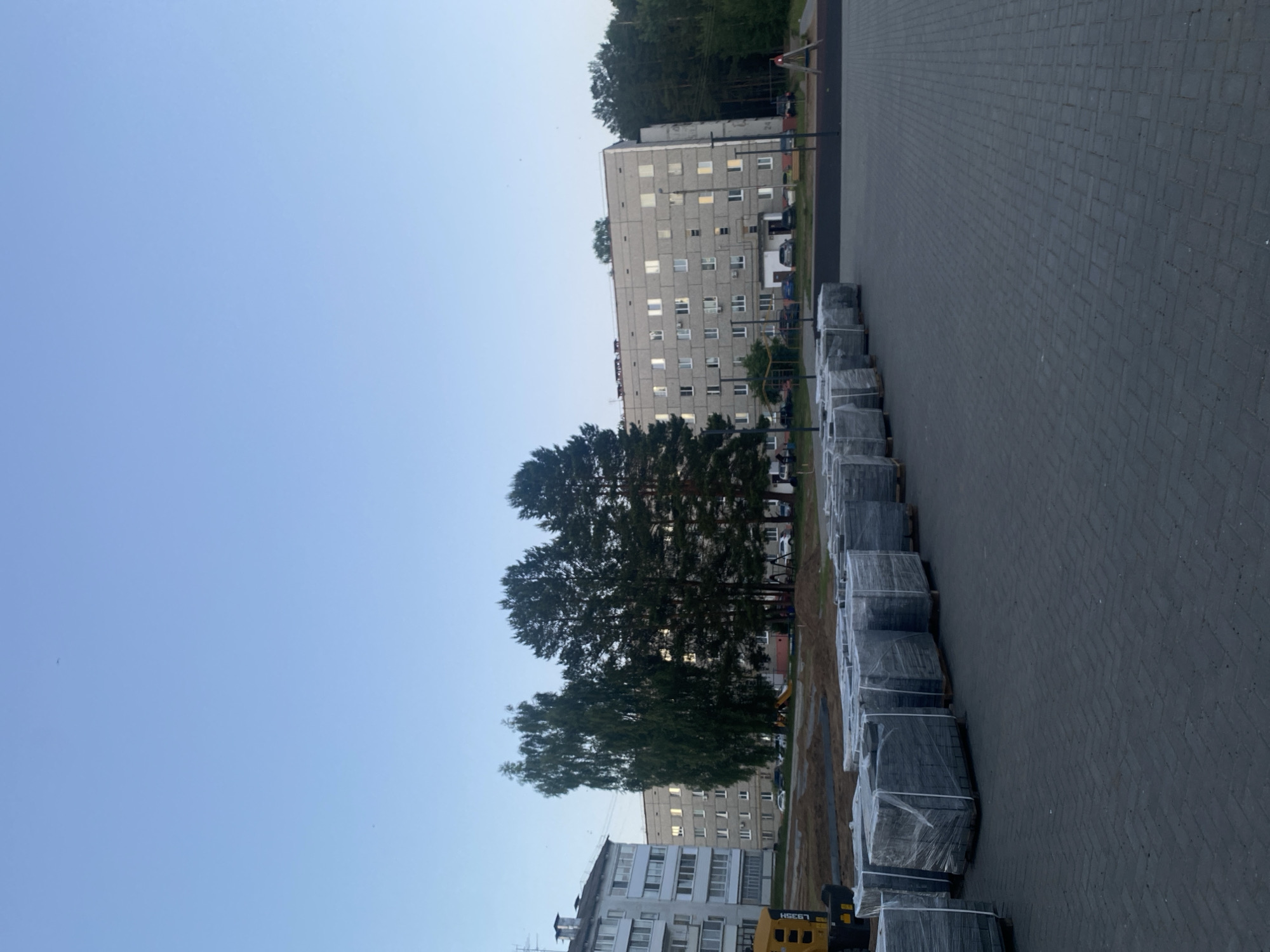 The image depicts a residential scene in Noviy, Russia, characterized by urban development materials and architecture. The foreground features a newly laid dark grey interlocking paver surface, extending from the bottom of the frame and receding towards the mid-ground. Along the left side of this paved area, a linear arrangement of numerous stacks of grey paving stones or bricks, individually wrapped in clear plastic film and placed on wooden pallets, are visible.
In the mid-ground, a large, rectangular, multi-story apartment building, constructed from light grey panels or concrete, stands prominently. This building, approximately five to six stories high, features numerous uniformly spaced rectangular windows, some reflecting ambient light and others showing internal illumination. Directly in front of and partially obscuring this central building are several tall trees, including one with lighter bark (possibly a birch) and several darker coniferous trees. To the left of the central building, a portion of another multi-story apartment building with balconies is visible. A yellow piece of construction equipment is partially visible at the bottom left, near the base of this building.
The area immediately surrounding the buildings and trees appears to be disturbed earth or an active construction site. In the far background on the right, a dense forest line is visible. The sky overhead is a clear, light blue, with a subtle gradient to a slightly darker shade higher up. Two small, dark specks, possibly birds, are discernible in the upper sky. No people are clearly visible within the frame.