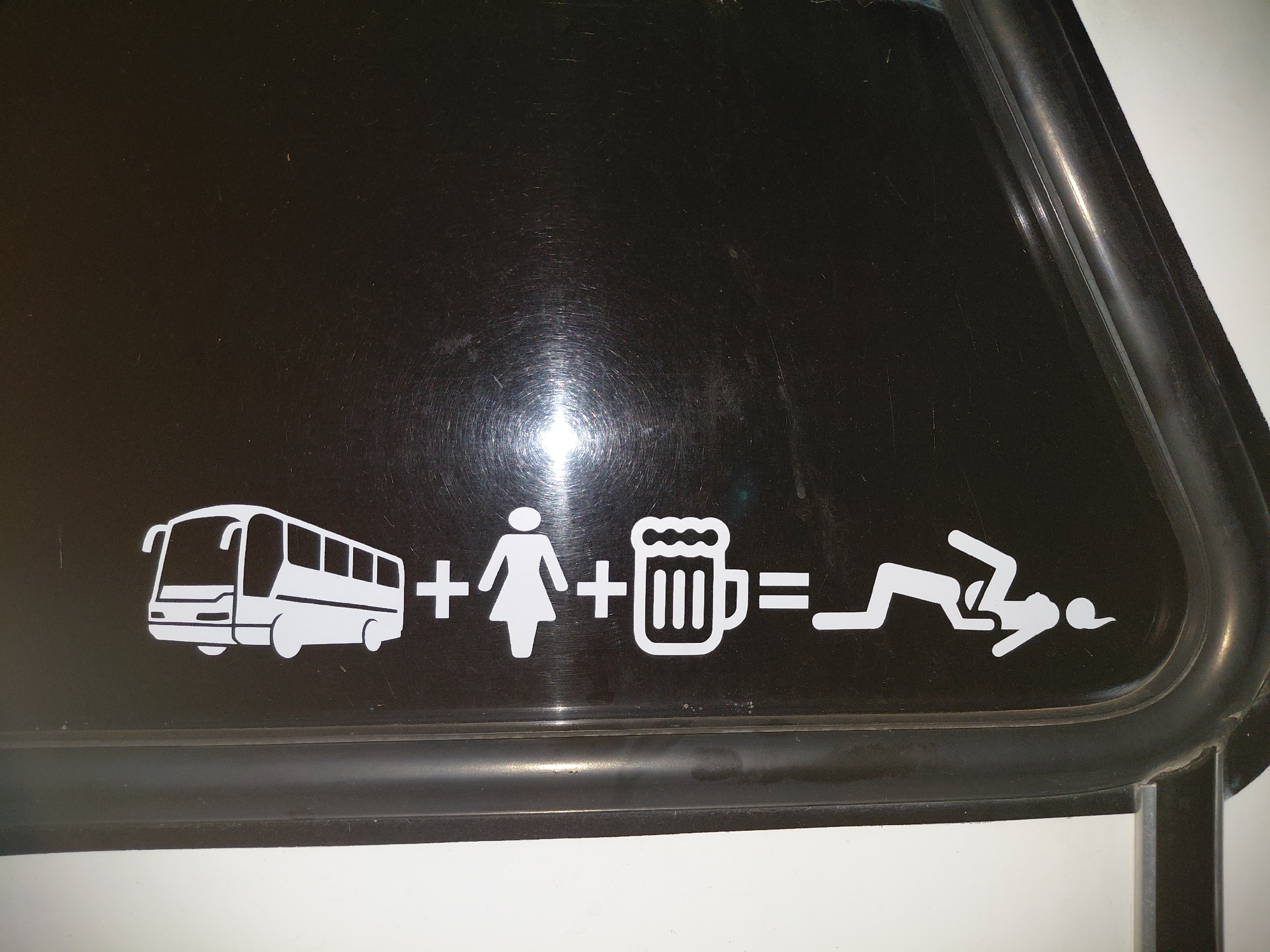 The image captures a close-up of a white pictogram decal affixed to a dark, glossy surface, likely a tinted vehicle window or body panel, in Santa Ana, Venezuela. A black rubber seal or trim frames the bottom and right edges of this surface.
The decal graphically illustrates a symbolic equation: a white outline of a passenger bus, followed by a plus sign (+), then a stylized white silhouette of a standing female figure, another plus sign (+), a white outline of a frothy beer mug, an equals sign (=), and finally, a white pictogram of a person lying prone on the ground, seemingly passed out or incapacitated.
The overall scene presents a humorous or light-hearted cautionary message, suggesting that the combination of a bus trip, female company, and beer can lead to someone becoming intoxicated to the point of collapse. The glossy dark surface prominently reflects a bright light source, creating a circular glare in the upper-left-center of the image. No visible text is present, only the symbolic pictograms and mathematical operators.