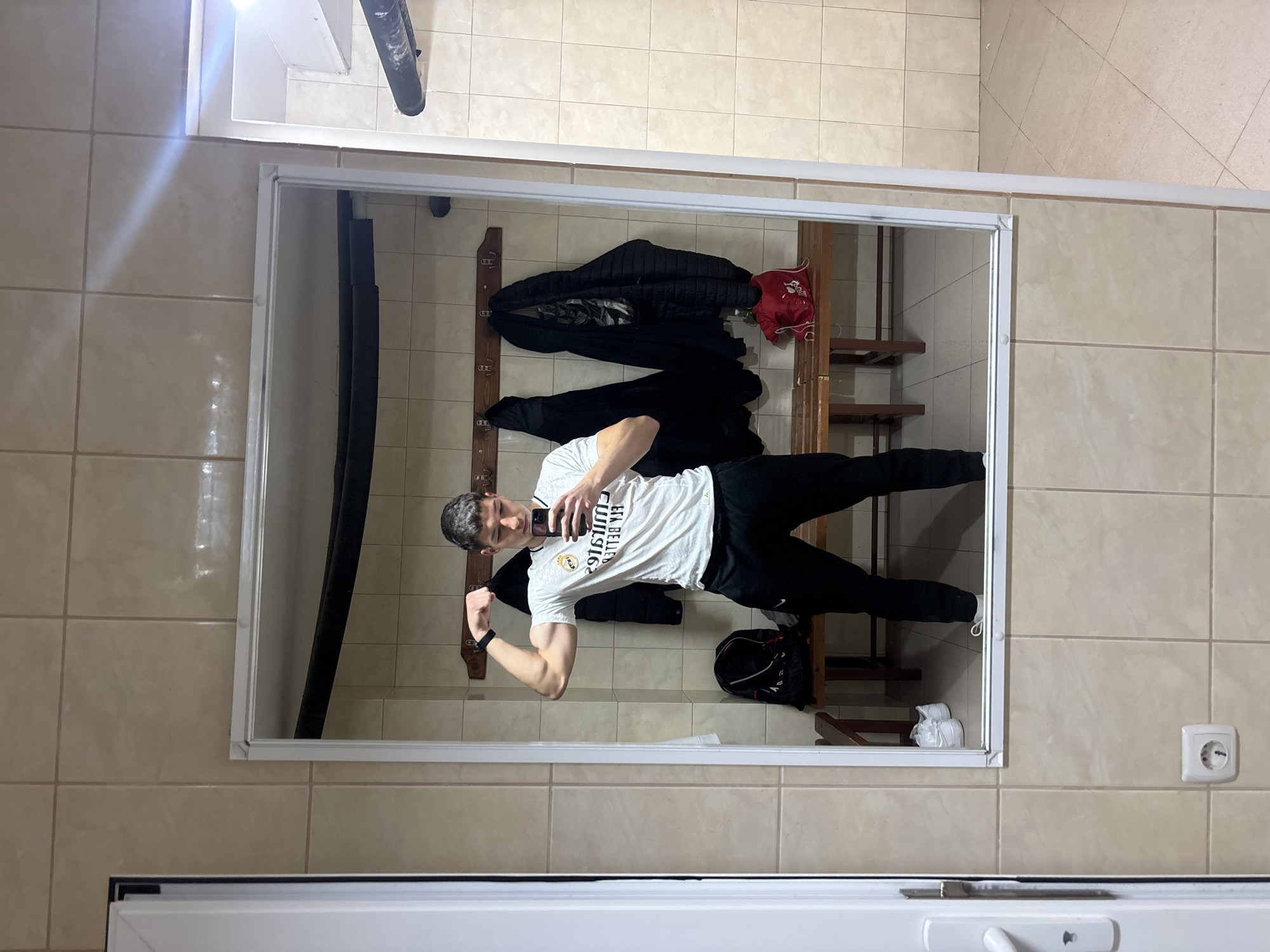 The image shows a man posing in front of a mirror in what appears to be a locker room or changing area. He is flexing his bicep and holding a phone in his other hand, likely taking a selfie. The room has light beige tiled walls. A coat rack with clothes and a bench are visible behind him in the mirror's reflection. An electrical outlet is present on the wall near the bottom right of the frame. There is no information that indicates the location in the city of Zlatograd, Bulgaria.