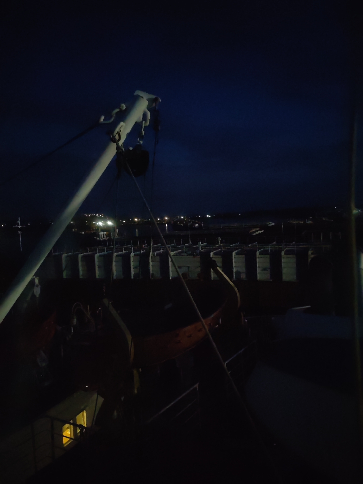 The image depicts a nighttime scene from what appears to be a boat or ship, possibly docked in Rybinsk, Russia. In the foreground, a white, angled metal pole is visible, along with dark equipment and an orange life raft. The background features a cityscape along the water, illuminated by scattered lights. There is no evidence of people in the image. The environment is dark, suggesting nighttime with limited artificial illumination.