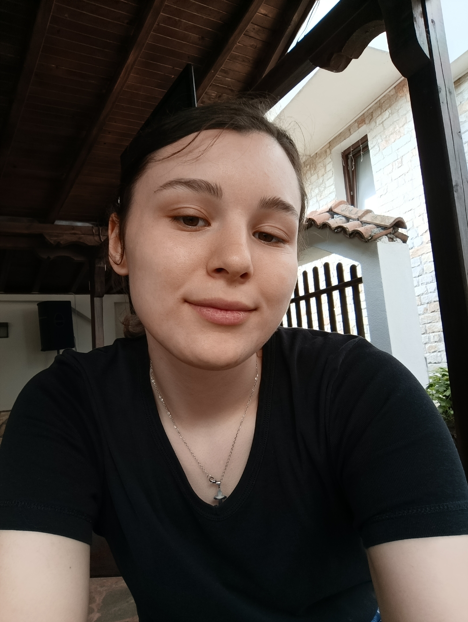 A young woman with brown hair and brown eyes is captured in a selfie taken outdoors. She is wearing a black v-neck t-shirt and a silver necklace with a cross pendant. Her left ear is visible, and she has a subtle smile. The background reveals a wooden-beamed roof structure overhead. To the left, a black speaker is mounted on a wall. Further back, a building with white brickwork and a tiled roof is visible, along with a dark metal fence. The overall setting suggests an outdoor patio or courtyard area, potentially in a residential or recreational space. The image does not contain explicit information about the city of Hadzhidimovo, Bulgaria.