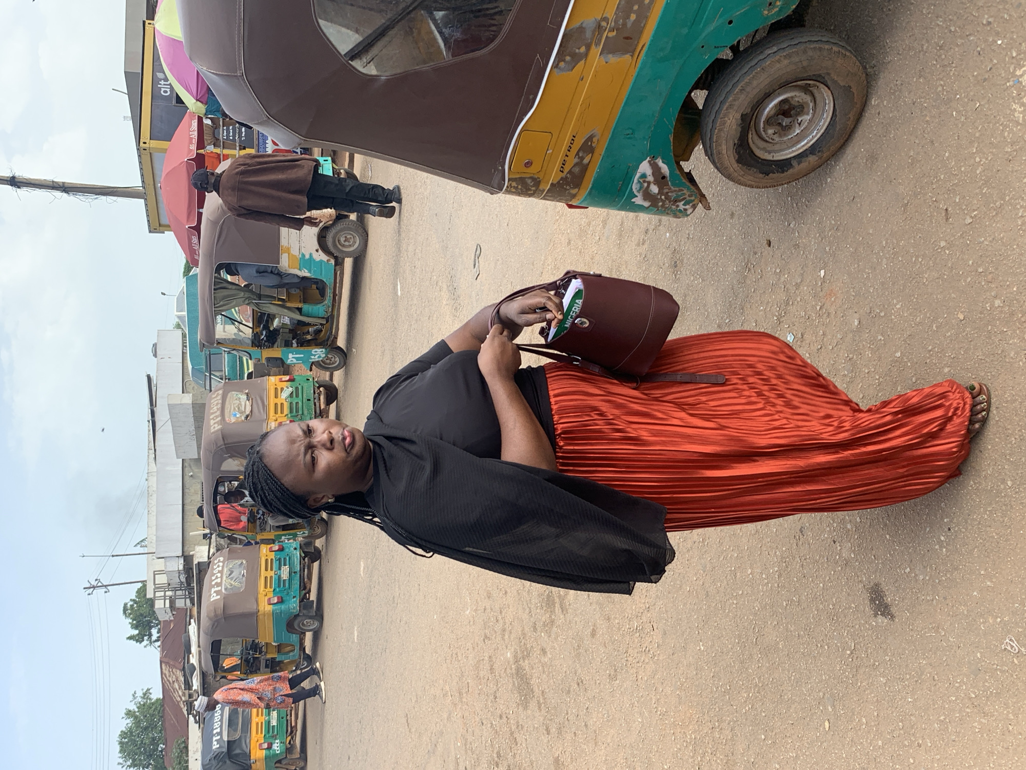 The image depicts a street scene in Gura Top, Nigeria, featuring a woman in the foreground wearing a black shawl and an orange pleated skirt, holding a dark red purse. Behind her, several auto rickshaws, known locally as "keke napeps," are parked or in motion. One keke napep is partially visible on the right side of the frame, predominantly yellow and green with "PETROL" printed on its side. Further back, a line of keke napeps stretches across the scene. The environment appears to be an open, dusty area, likely a transportation hub or marketplace, with some buildings and umbrellas visible in the background. The sky is overcast.