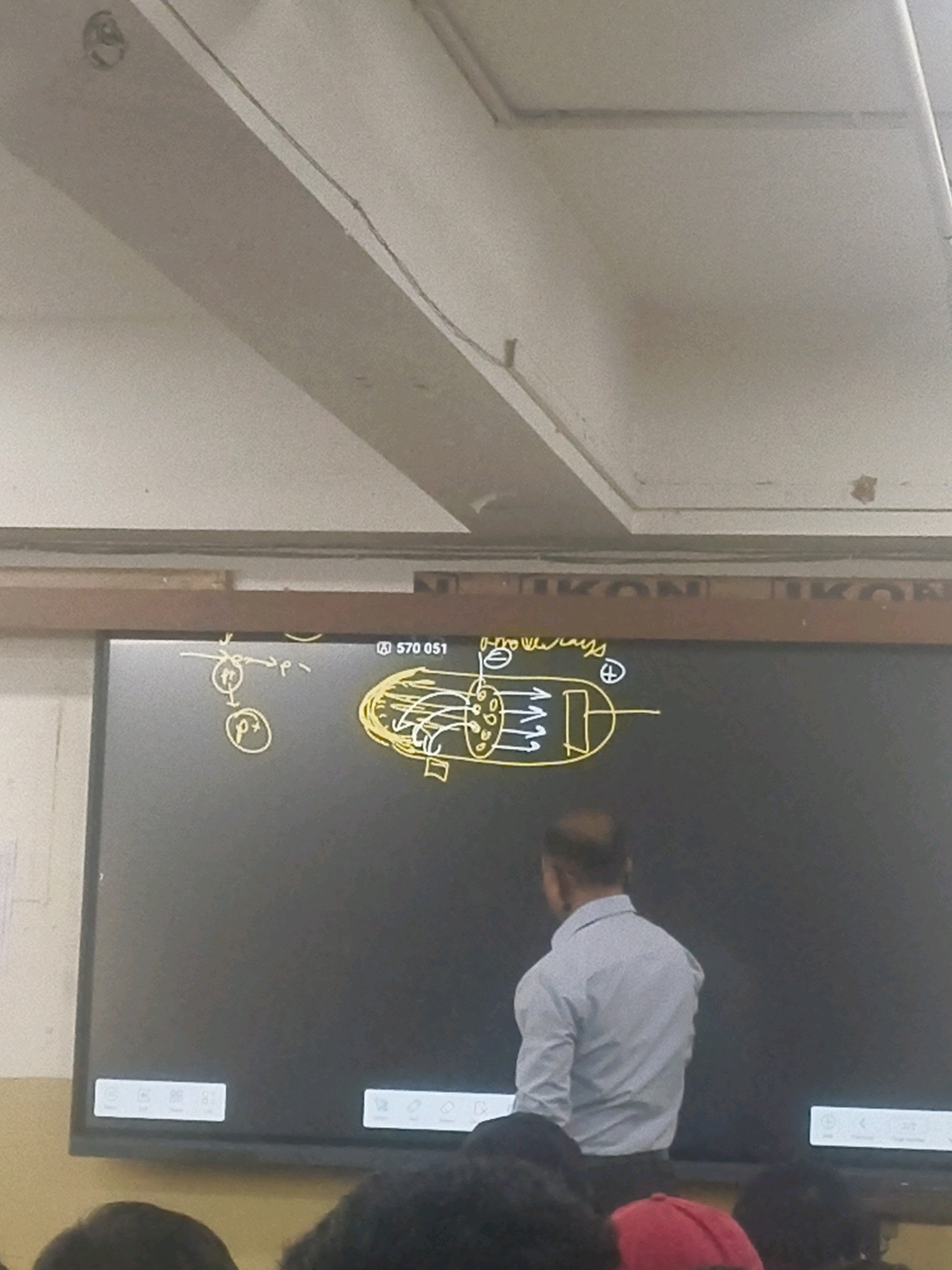 In a classroom setting, likely in Kota, India, a digital whiteboard displays a scientific diagram of an electronic component, possibly a vacuum tube or a similar device, with symbols representing electrical charges and electron flow. The drawing is in yellow, with a plus sign and a minus sign prominently shown. To the left of the diagram, smaller circular diagrams depict atomic particles, labeled with "p+" for a proton and "e-" for an electron, indicating a discussion related to subatomic particles or their behavior. A man, presumably a teacher, stands facing the board, his back mostly to the camera, wearing a light-colored collared shirt. The lower part of the image shows the heads of students, their faces obscured, suggesting they are seated and attentive. The environment appears to be a typical educational institution with a large screen display and the subtle architectural features of a ceiling.