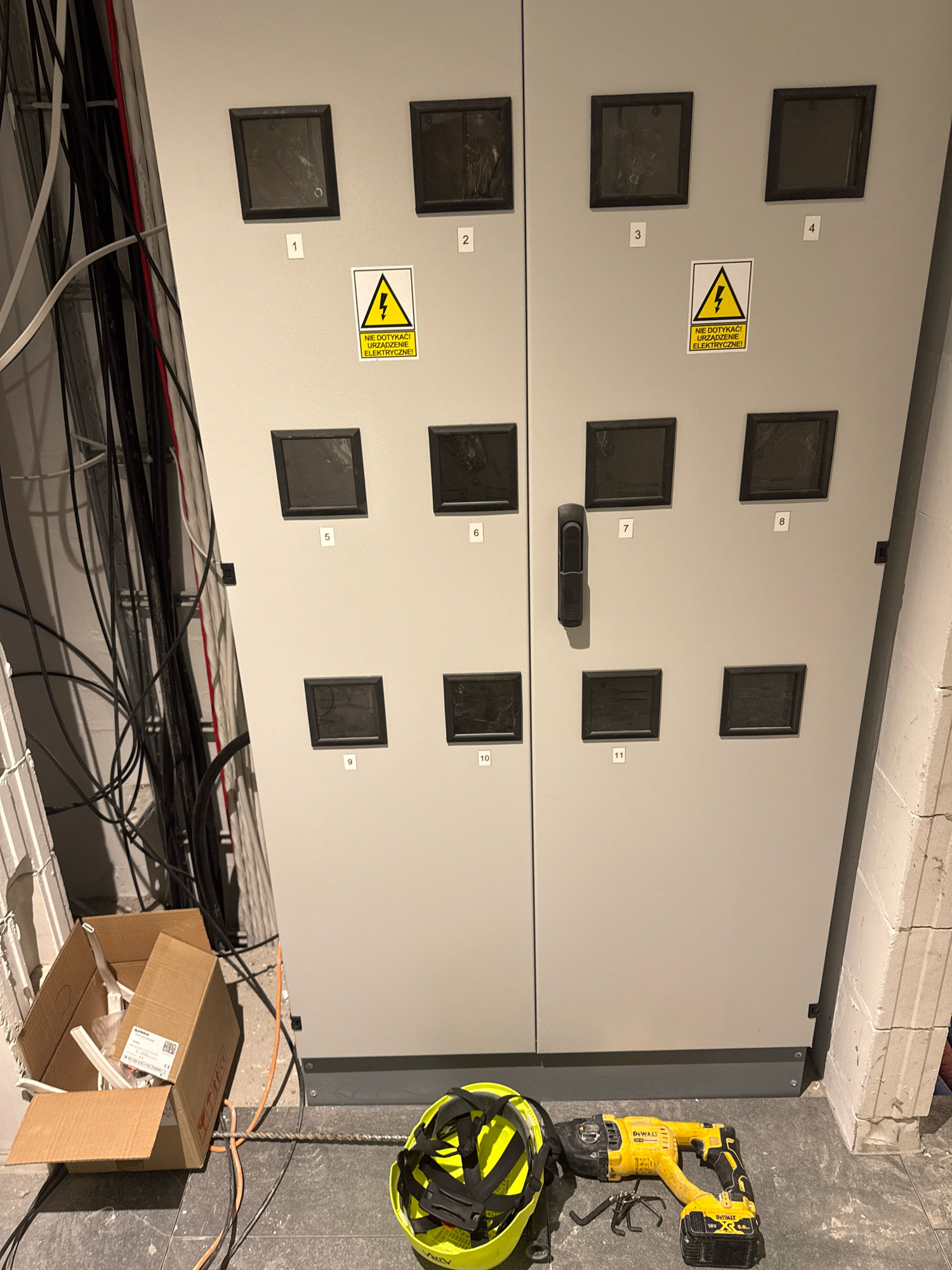 In a dimly lit room, likely a utility or electrical closet, a large, light gray electrical panel dominates the frame. The panel is divided into two vertical sections, each featuring a row of four dark, recessed squares, presumably access panels or windows. Below these, on the left section, are numbered labels '1' through '4', and on the right section, '5' through '8'. The center of the right section has a black vertical handle. Below the top row of squares on the left, there is a yellow warning sticker with a black lightning bolt symbol and Polish text reading "NIE DOTYKAĆ! URZĄDZENIE ELEKTRYCZNE!" (Do not touch! Electrical device!). A similar sticker is located below square number '4' on the right side of the panel. Further down, numbered labels '9', '10', '11', and '12' are visible below squares on the lower portion of the panel.
To the left of the electrical panel, a collection of black and red electrical cables are neatly organized and run vertically along the wall, some secured with white zip ties. In the foreground, on the floor, is an open cardboard box containing white electrical components. Beside the box lies a bright yellow DeWalt hammer drill with a black battery pack. Adjacent to the drill is a yellow safety helmet with black straps. The floor is a dark gray tiled surface. The scene suggests an ongoing electrical installation or maintenance project. The location is identified as Nowa Sól, Poland.