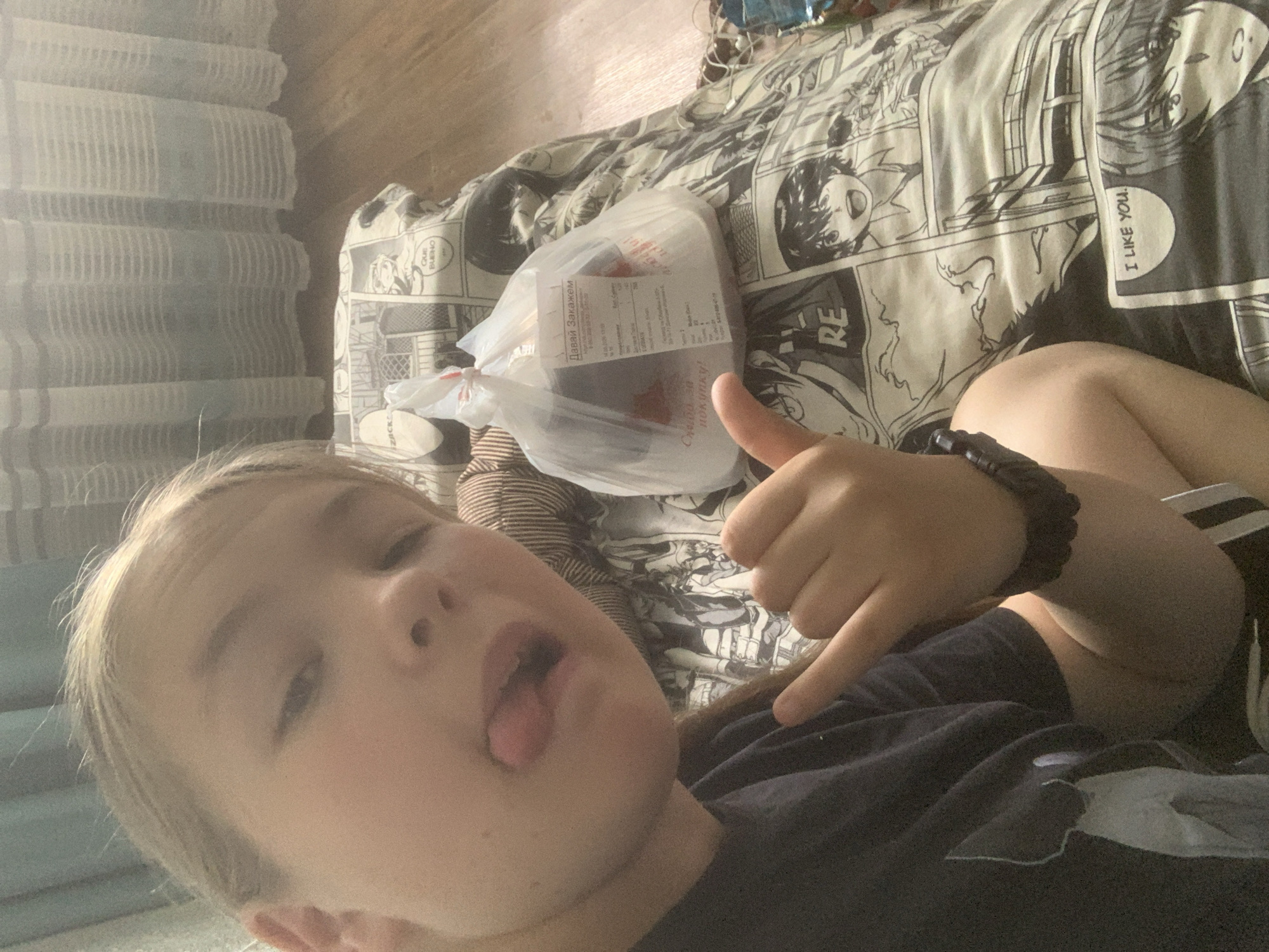 A young child with blonde hair is lying down and making a funny face, sticking their tongue out. They are wearing a dark t-shirt and a black beaded bracelet. To the right of the child is a clear plastic bag containing food, with a receipt visible inside. The bag is sitting on a bed with a black and white anime-themed duvet cover. The duvet features various manga panels and characters, with some visible text in English, such as "I LIKE YOU." and "RE". In the background to the left, a stack of white objects, possibly plates or containers, are visible. The scene appears to be indoors, likely in a bedroom, during the daytime, suggested by the lighting. The overall mood is playful and casual. Visible text on the receipt includes "Давай Закажем" (Let's Order) and "Слибо за покупку!" (Thanks for the purchase!).