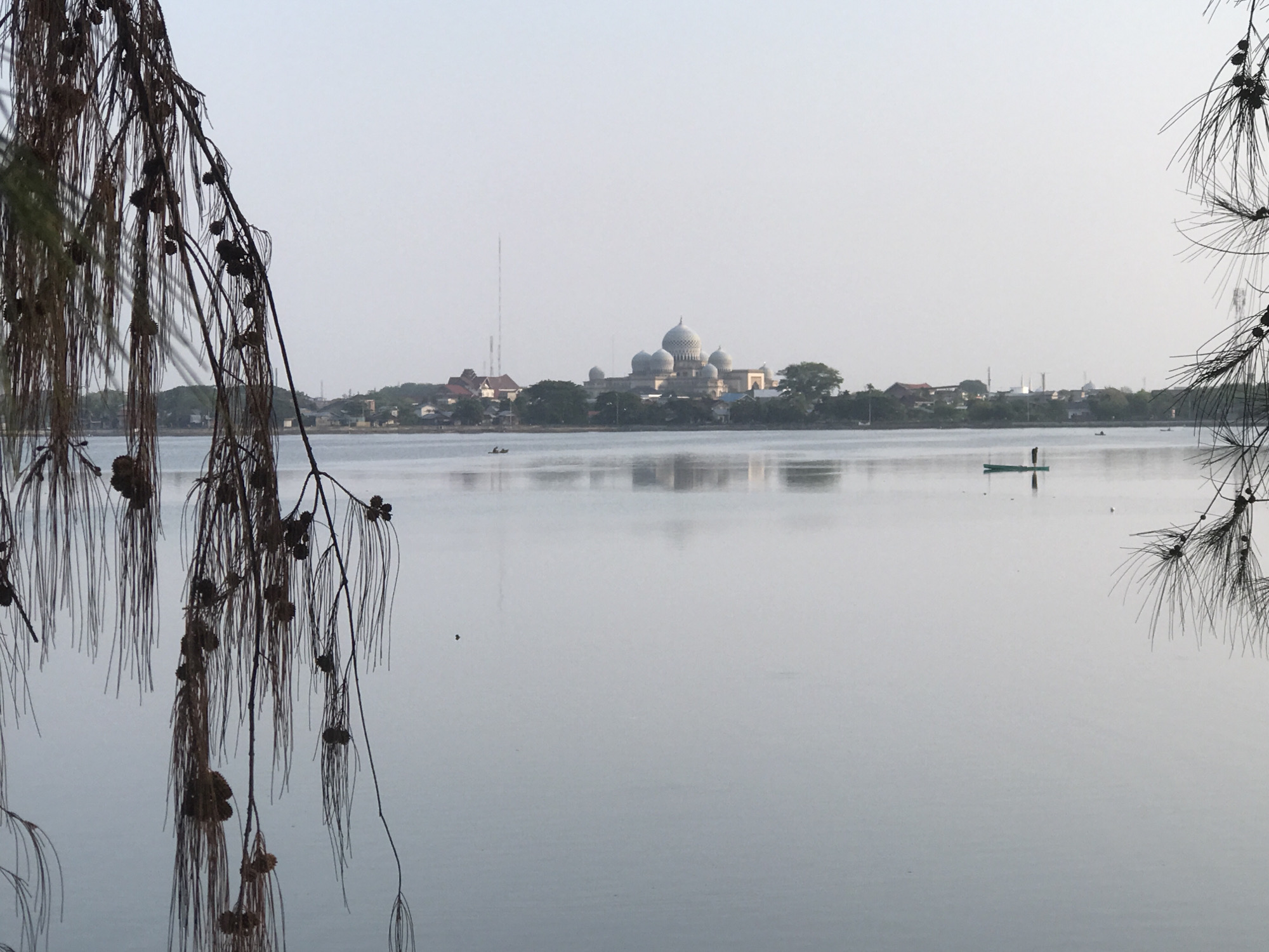 The photograph captures a serene view of a calm body of water in Lhokseumawe, Indonesia, with a prominent mosque featuring multiple domes in the background. A person stands on a narrow, green boat on the right side of the frame. Another small boat appears further in the distance. The foreground is marked by the branches and drooping needles of a tree on both sides, with visible pine cones. The water reflects the buildings and sky.