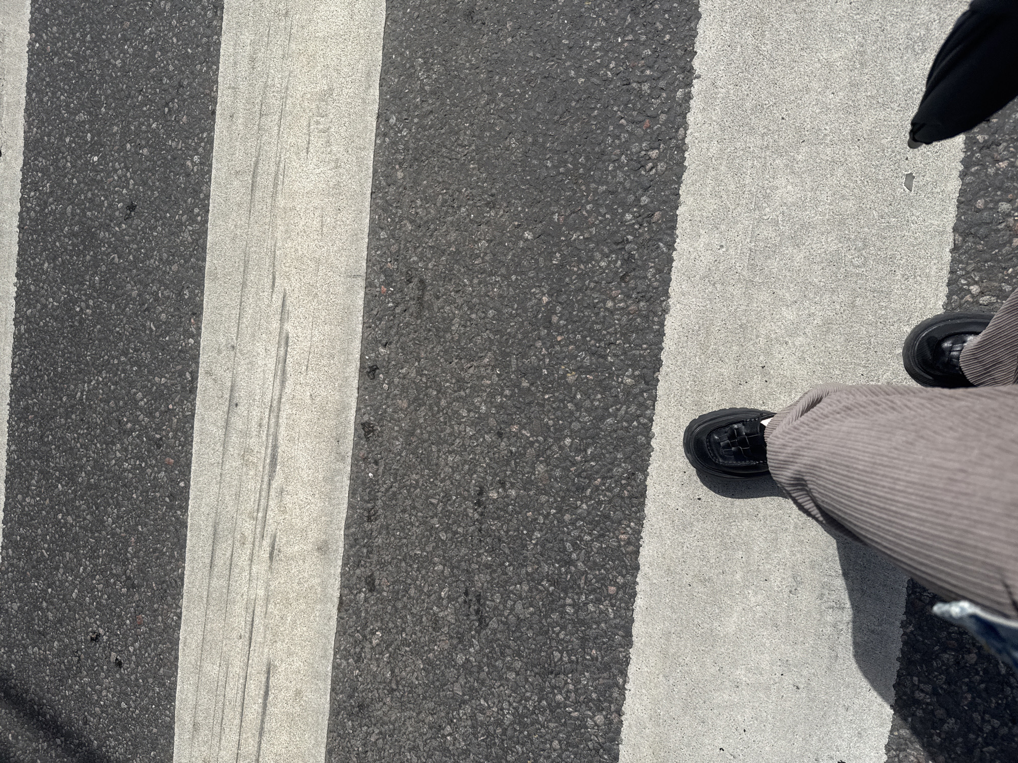 The image shows a close-up view of a person's legs and feet standing on a pedestrian crossing. The crossing is marked with white stripes on an asphalt road. The person is wearing brown corduroy pants and black chunky-soled shoes. A shadow is cast on the white stripe to the left of the person's legs. The scene is outdoors, under daylight. There are no visible landmarks or signs to indicate a specific location in Moscow, Russia.