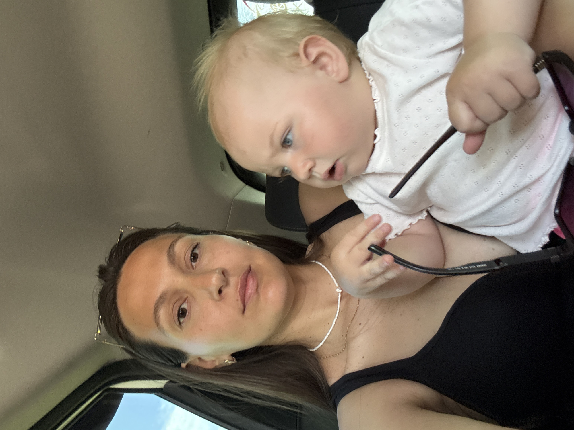 In the city of Chaikovsky, Russia, a woman and a baby are seen inside a vehicle, likely a car, given the view of the car's interior and the presence of a window with a sky visible outside. The woman, with her long, dark hair and light complexion, is wearing a black top and a delicate white beaded necklace. She is holding a pair of sunglasses. The baby, with fair hair and blue eyes, is dressed in a light pink shirt with a subtle patterned texture. The baby is looking to the side with an open mouth, while the woman is looking directly at the camera. The interior of the car appears to be dimly lit, with the natural light coming from the window. The overall scene suggests a candid moment captured while traveling.
