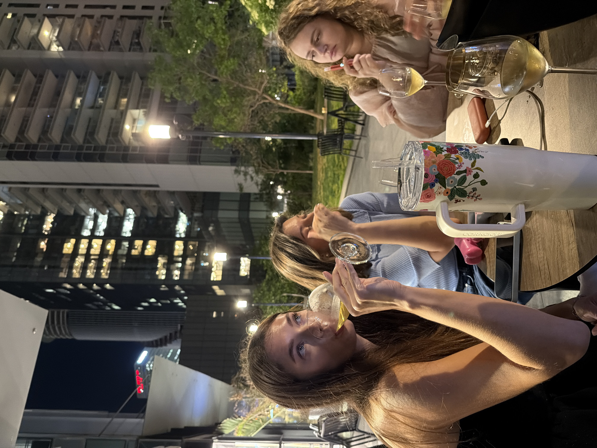 The image shows three women seated at a table outdoors at night. They are drinking white wine. A large building with many windows is in the background, suggesting an urban environment. In the foreground, there is a decorated water bottle on the table. The location appears to be Tel Aviv, Israel, given the style of the buildings and the general ambience. The women are engaged in conversation and enjoyment of the evening. One woman has a device resembling an e-cigarette in her hand. They are seated on a patio, possibly part of a restaurant or bar, suggested by outdoor furniture visible in the background.