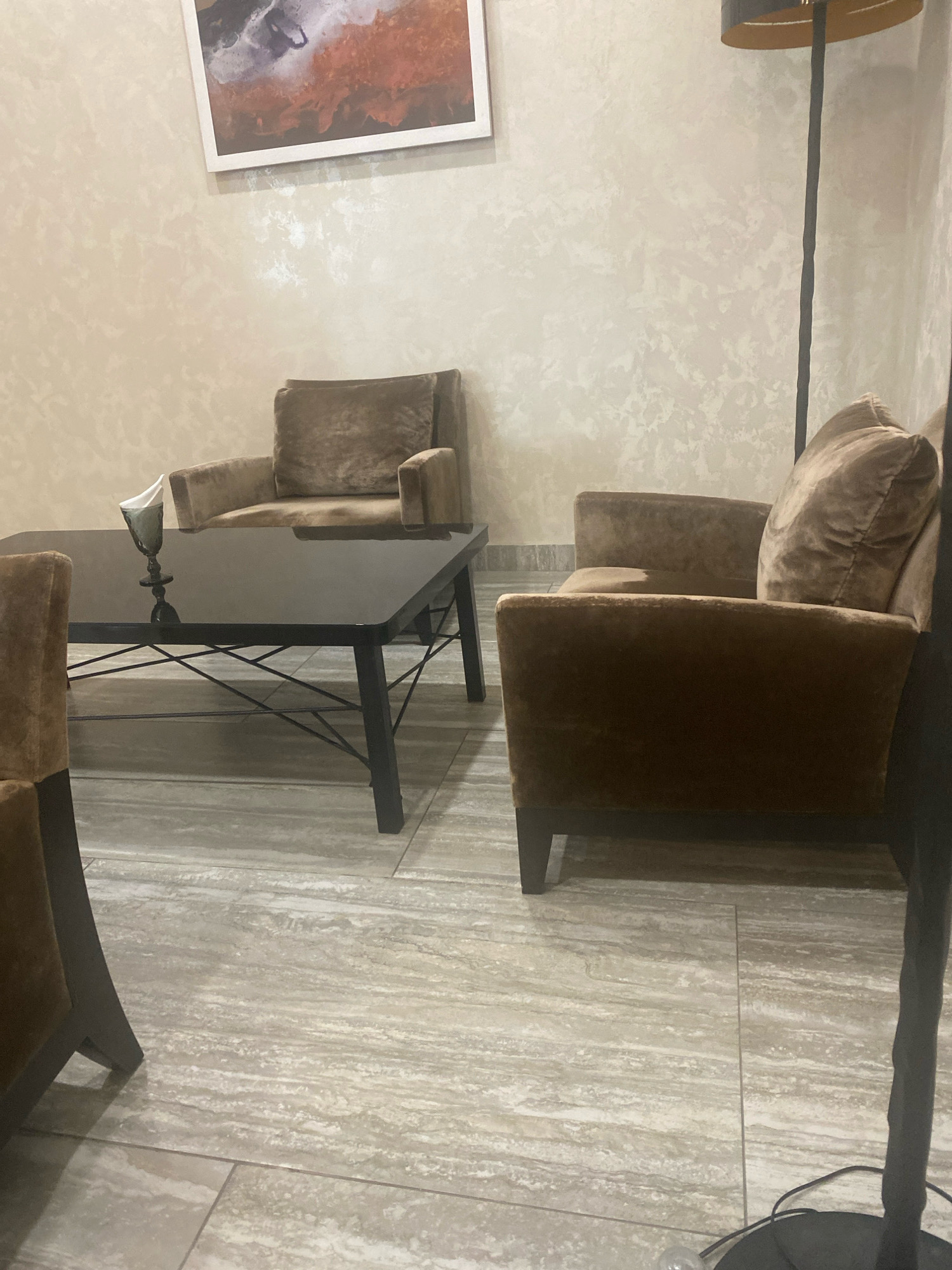 The image depicts an interior scene likely in a cafe or lounge area in Minsk, Belarus, featuring a contemporary seating arrangement. In the foreground, a dark, rectangular coffee table with a glossy surface is centrally positioned. On the table, a decorative glass or vase with a white napkin is visible. Two plush, brown armchairs are present in the scene. One armchair is fully visible on the right side of the frame, while another is partially seen on the left. A third armchair is positioned behind the coffee table, with its back and headrest facing the camera. The walls are adorned with a textured wallpaper in a light, neutral tone. A framed piece of art, featuring abstract shapes in earthy colors, hangs on the wall above the armchair in the background. A tall, dark floor lamp stands to the right of the frame, with its shade and a portion of the pole visible. The floor is tiled with large, light-colored tiles that have a natural stone-like pattern. The overall ambiance suggests a comfortable and stylish waiting or lounging area.