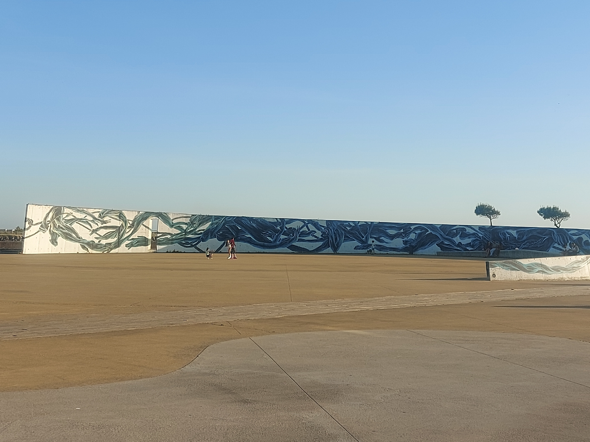 The image shows a public space in Figueira da Foz, Portugal, with a long, low wall featuring a mural of blue and white wave-like patterns. The sky is clear and blue. Two small trees are visible behind the right side of the wall. Several individuals are scattered throughout the scene: a person is walking in front of the mural, and other people are near the right side of the wall. The ground is predominantly a flat, paved surface with areas of different textures and colors.