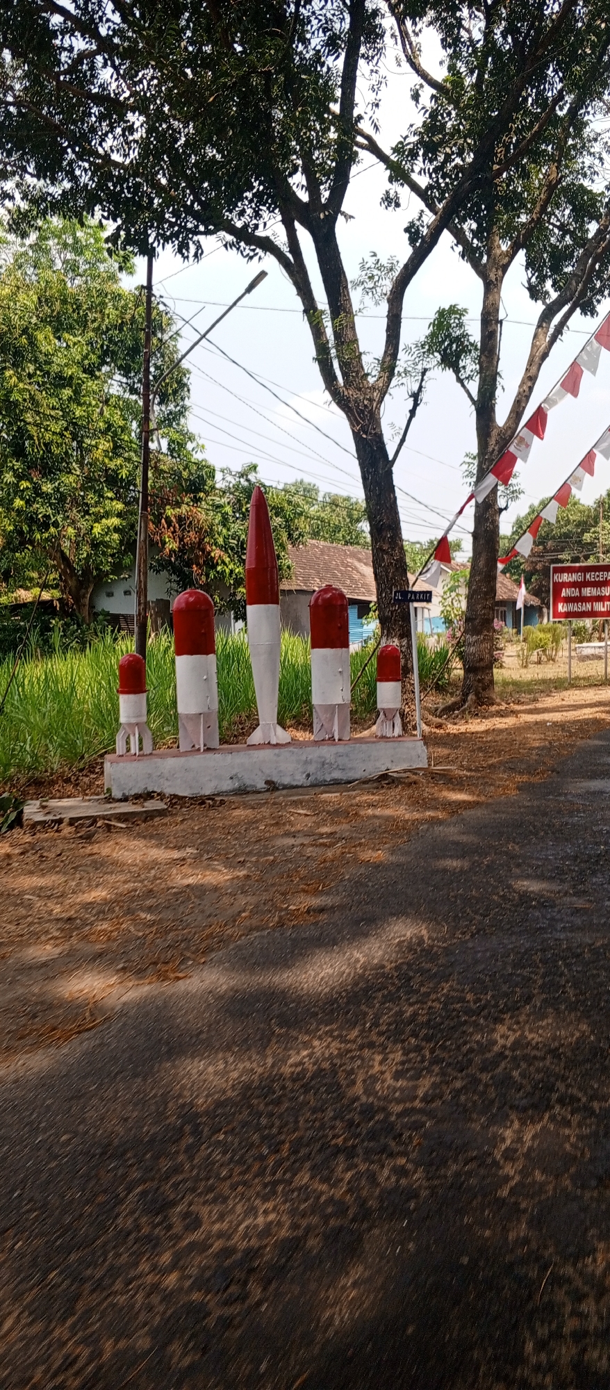 The image depicts a roadside scene in Maospati, Indonesia, likely near an entrance to a military area given the signage. In the foreground, a paved road transitions from a dirt and leaf-covered shoulder. Along the side of the road, there is a concrete barrier or planter box. Mounted on this barrier are several decorative objects resembling rockets or missiles, painted in alternating red and white bands, with one larger, pointed missile-shaped object standing taller than the others. These are interspersed with smaller, cylindrical red and white markers. Behind the barrier, tall green grass grows, and further back, a building with a brown tiled roof is visible, partially obscured by trees. A street sign reading "JL. PARKIT" is mounted on a pole. Across the street, more trees are present, and red and white Indonesian flags are strung in a festive manner. A sign with Indonesian text, likely a warning or informational notice, is posted on a pole. The overall environment suggests a sunny day in a semi-rural or suburban area, possibly marking an official or commemorative site. No people are visible in the image.