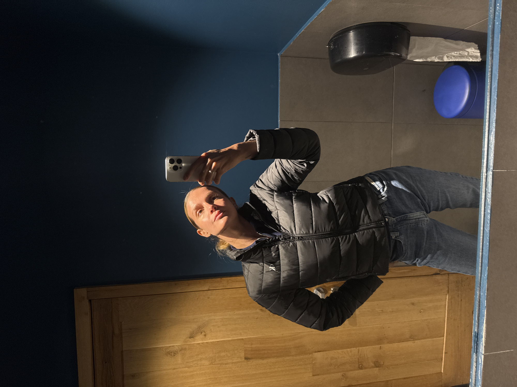 A person is taking a selfie in a bathroom. The person is wearing a black puffer jacket and blue jeans. The bathroom has a blue wall, a wooden cabinet, and a mirror. On the wall near the mirror are a black container, toilet paper, and a blue container. The city of Tbilisi, Georgia is not discernible from the image.