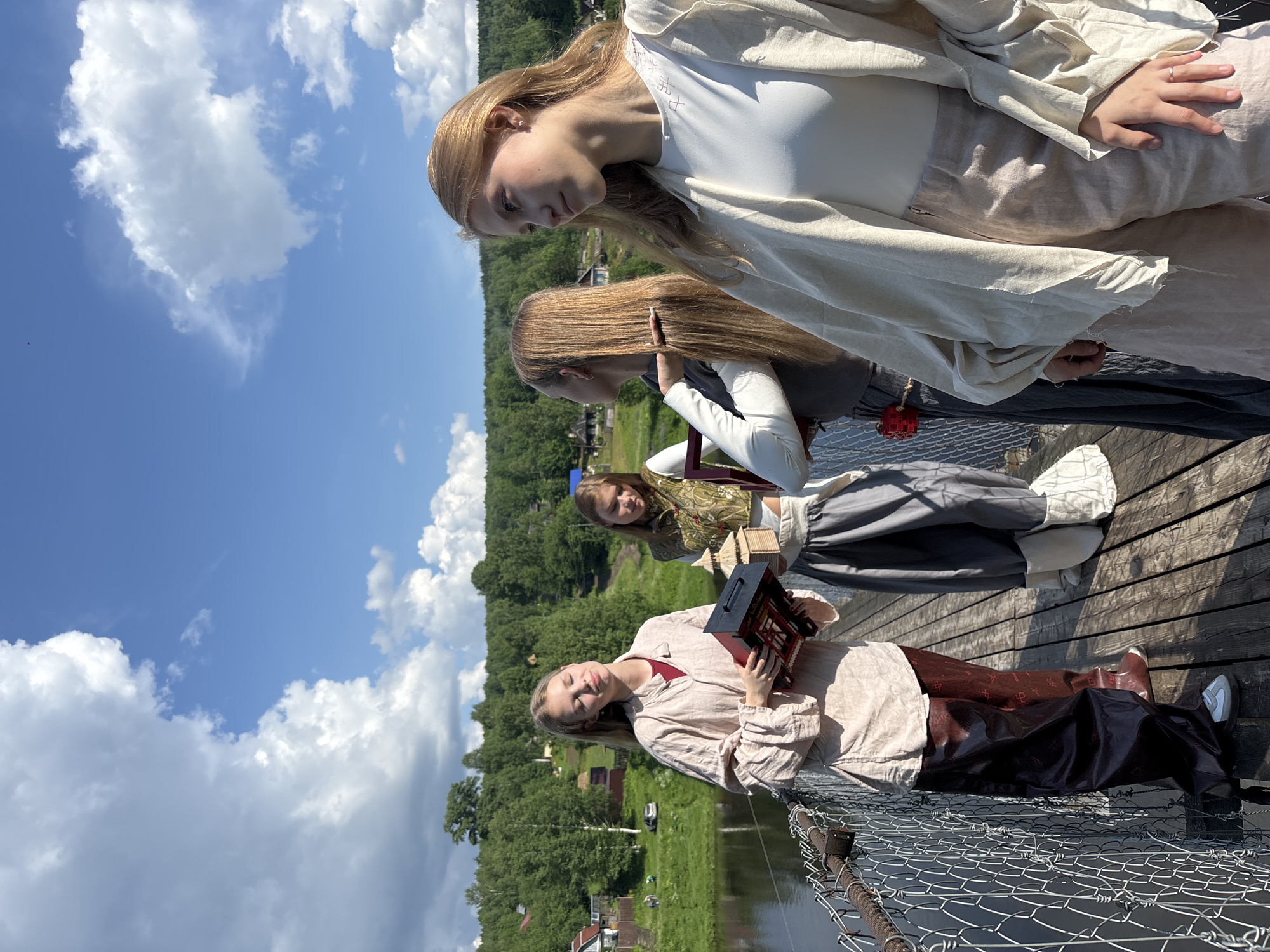 Four young women stand on a wooden suspension bridge on a sunny day. The bridge spans a body of water and is flanked by a chain-link fence. In the background, a lush green forest lines the hillsides, with scattered houses and buildings visible. The sky is a bright blue with fluffy white clouds.
The woman closest to the viewer on the right is wearing a light-colored, loose-fitting outer garment over a white top and light-colored pants. Her blonde hair is down, and she is looking to her right, with her left hand resting on her lap.
To her left, another woman with blonde hair, also down, is wearing a dark long-sleeved shirt and a gray skirt. She is looking towards the woman to her left. A red object is attached to her belt.
In the center, a woman with blonde hair tied back is wearing a vest over a white top and a gray skirt. She is holding a small, intricately designed wooden structure.
On the far left, a woman with blonde hair is wearing a light-colored tunic and dark, wide-legged pants with a patterned print. She is holding a red and black box. She is looking down with her eyes closed.
The scene appears to be set in the city of Gubakha, Russia, given the style of the wooden bridge and the surrounding architecture.