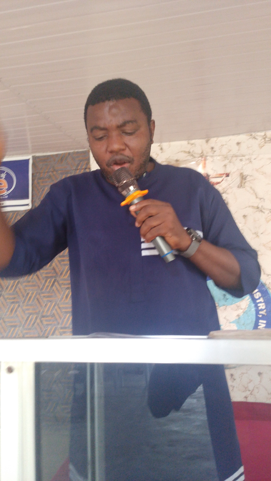 A man with dark skin and short black hair is singing into a silver microphone with an orange windscreen. He is wearing a dark blue long-sleeved shirt. A watch is visible on his left wrist. He appears to be standing behind a white surface. In the background, there is a wall with a patterned design and a light-colored ceiling. There is also a blue and white circular logo visible on the wall. The overall environment appears to be indoors.