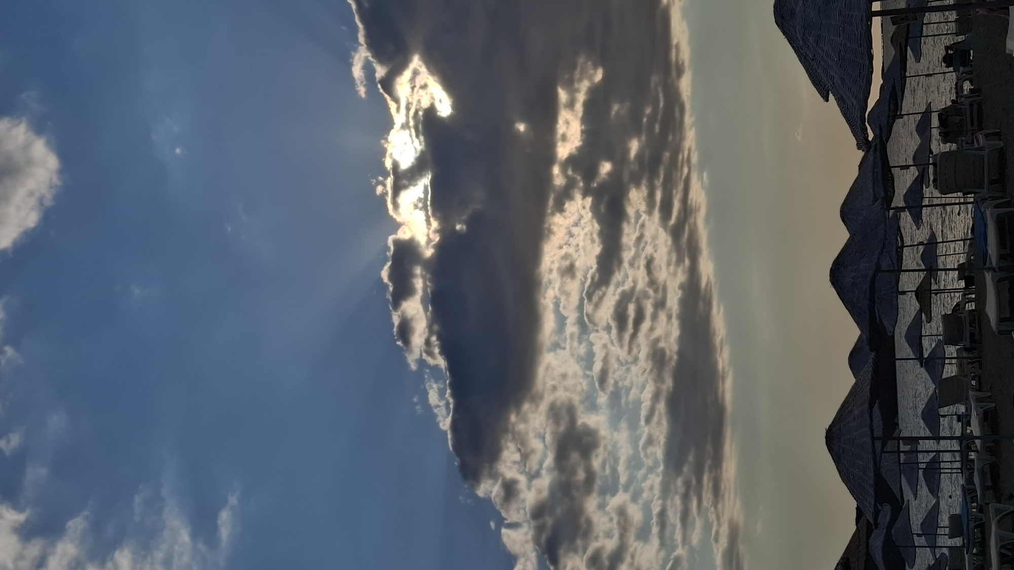 A vertical, low-angle shot captures a dramatic sunset sky over a beach in Foça, Turkey. The sun peeks through a band of dark clouds, casting ethereal rays downward. Below the sky, rows of thatched-roof beach umbrellas and empty lounge chairs are silhouetted against the calmer sea. The beach appears deserted.