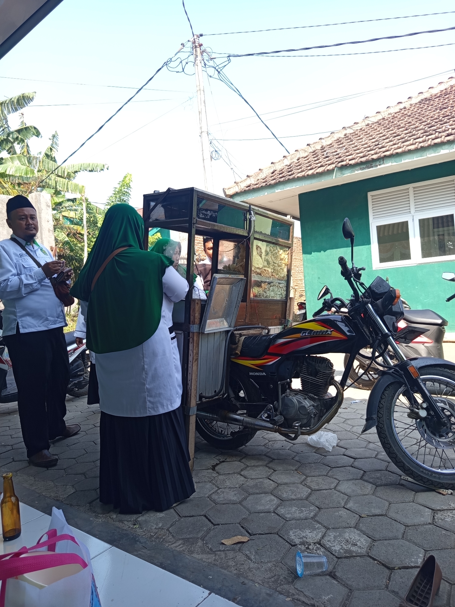 A group of people are gathered around a food cart on a paved street in Lumutan, Indonesia. A man wearing a white long-sleeved shirt, black pants, and a black cap stands on the left, holding a mobile phone. A woman in a green hijab and a white long-sleeved shirt with a black pleated skirt is to his right, facing the food cart. Another woman, also in a green hijab and white clothing, is visible behind the first woman, partially obscured. The food cart is a metal structure with glass panels displaying food items. A black motorcycle with red, yellow, and black decals is parked next to the cart, facing forward. In the background, a green building with white-slatted windows and a tiled roof is visible. The sky is clear and blue. Debris, including a plastic cup and a crumpled plastic bag, is scattered on the ground near the motorcycle. A glass bottle and a pink and white bag are on a white surface in the foreground.