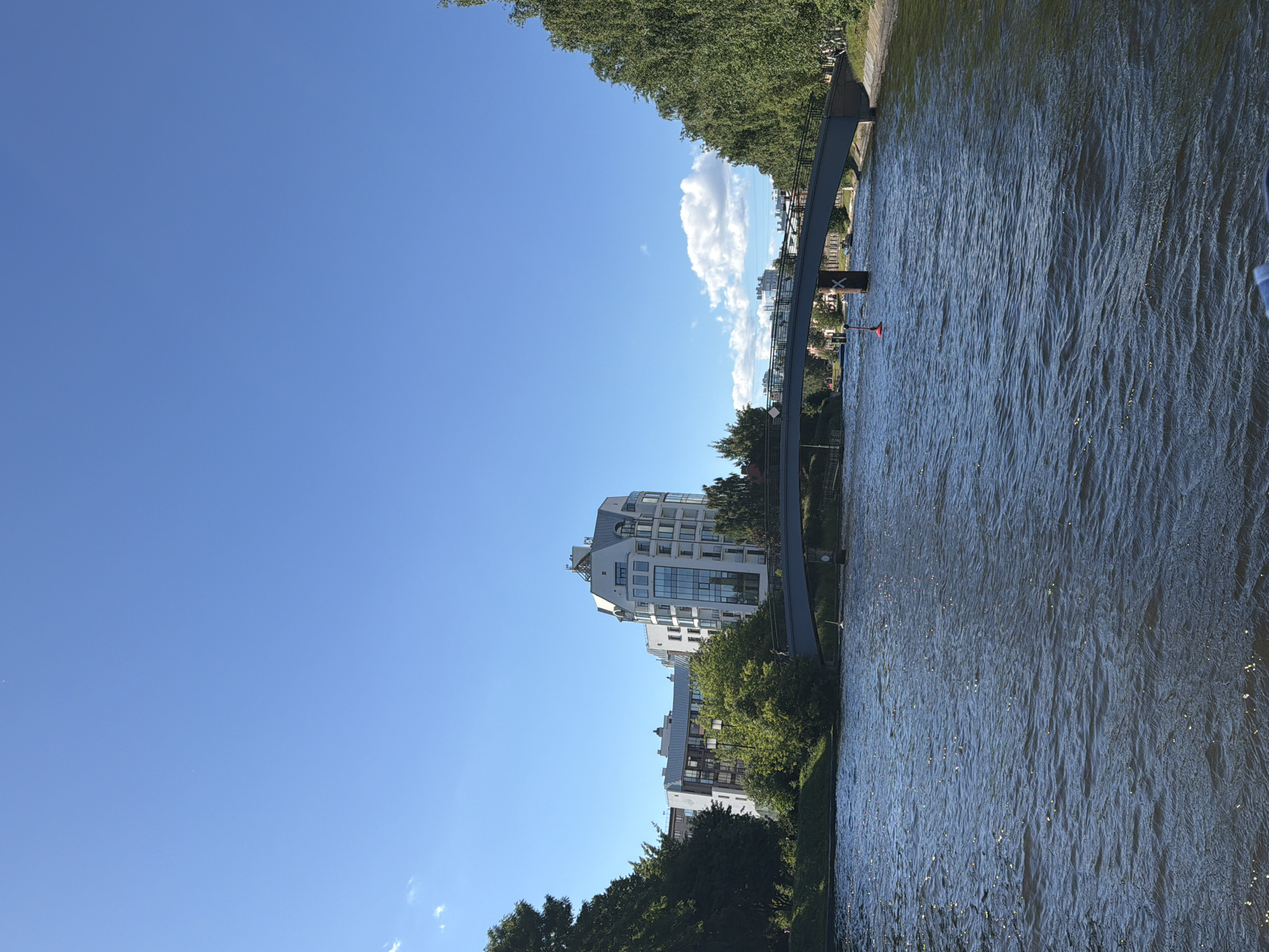 A wide, choppy river occupies the foreground, reflecting sunlight in numerous small glints. A grey metal bridge with a railing spans the river in the midground, connecting two tree-lined banks. On the left bank, several modern buildings with white facades and large windows are visible behind a dense growth of green trees. The tallest building has a curved, distinctive roof. On the right bank, a dense line of mature green trees forms a solid wall. The sky above is a clear, bright blue with a few wispy white clouds. The scene appears to be a daytime view of a city waterway.
