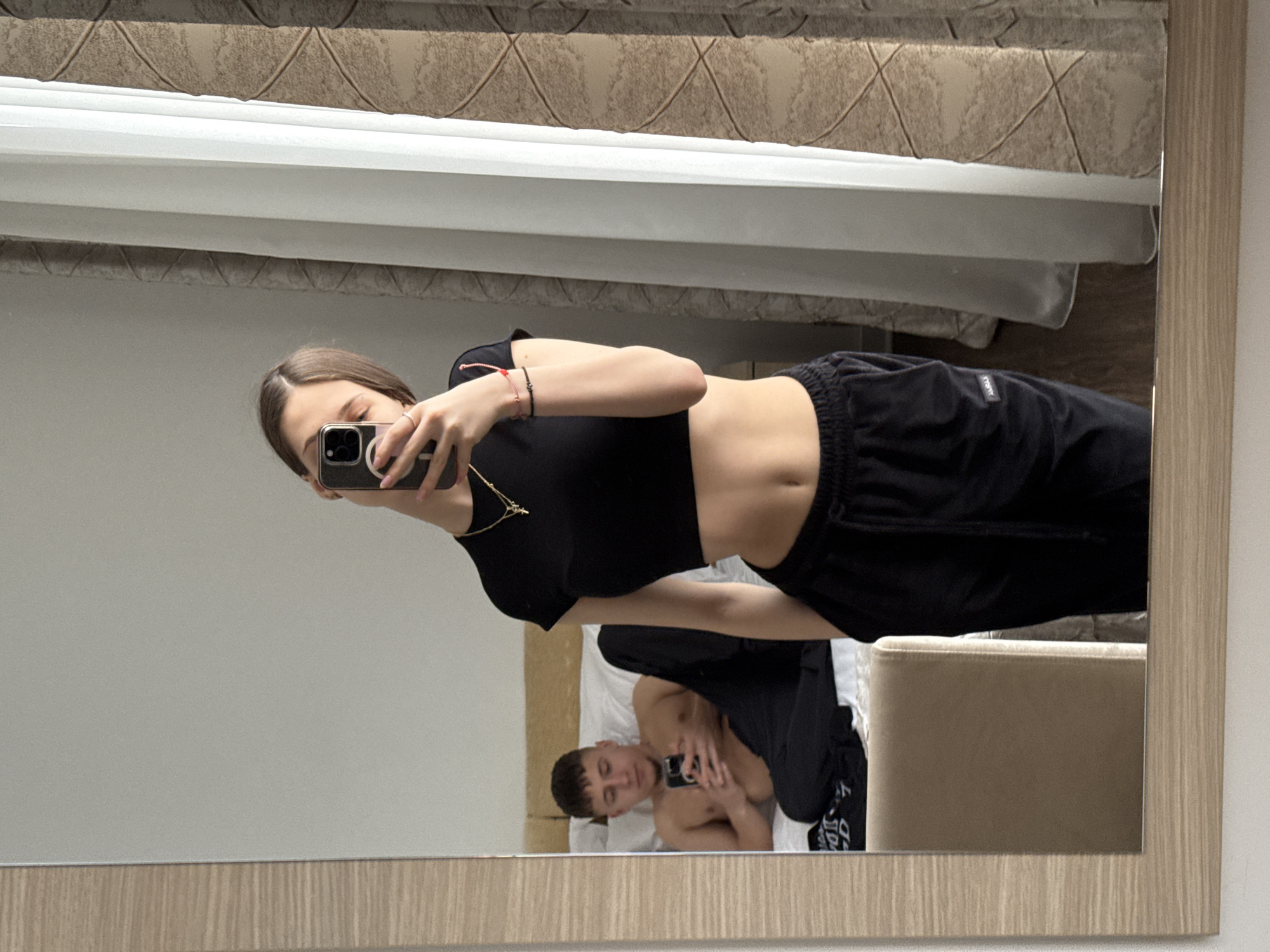 The image depicts an indoor scene, specifically a bedroom or hotel room, located within the Vozdovac Urban Municipality, Serbia. A large rectangular mirror with a light wood-grain frame occupies the left and central portion of the frame.
In the foreground of the mirror's reflection, a young adult female is visible from the chest down to the mid-thigh, taking a mirror selfie with a dark-colored smartphone. Her face is partially obscured by the phone. She wears a black short-sleeved crop top, exposing her midriff, and black drawstring sweatpants. Accessories include a gold-colored necklace with a small pendant, a ring on her right hand, and a red and black bracelet on her left wrist. Her hair is dark and straight, parted on the side. One hand holds the smartphone, while the other is in her sweatpants pocket.
In the background of the reflection, a young adult male is reclining on a bed with white bedding and multiple white pillows, propped against a light-colored headboard. He is bare-chested, wearing dark-colored pants. He holds a separate dark-colored smartphone with both hands and appears to be looking at its screen.
The room features plain light-colored walls. To the right of the mirror, a window with a sheer white curtain and a patterned beige/brown opaque curtain is visible, through which natural light enters. A portion of a light brown upholstered chair or furniture item is reflected in the lower left section of the mirror. The floor, visible in the lower right reflection, appears to be dark-colored.