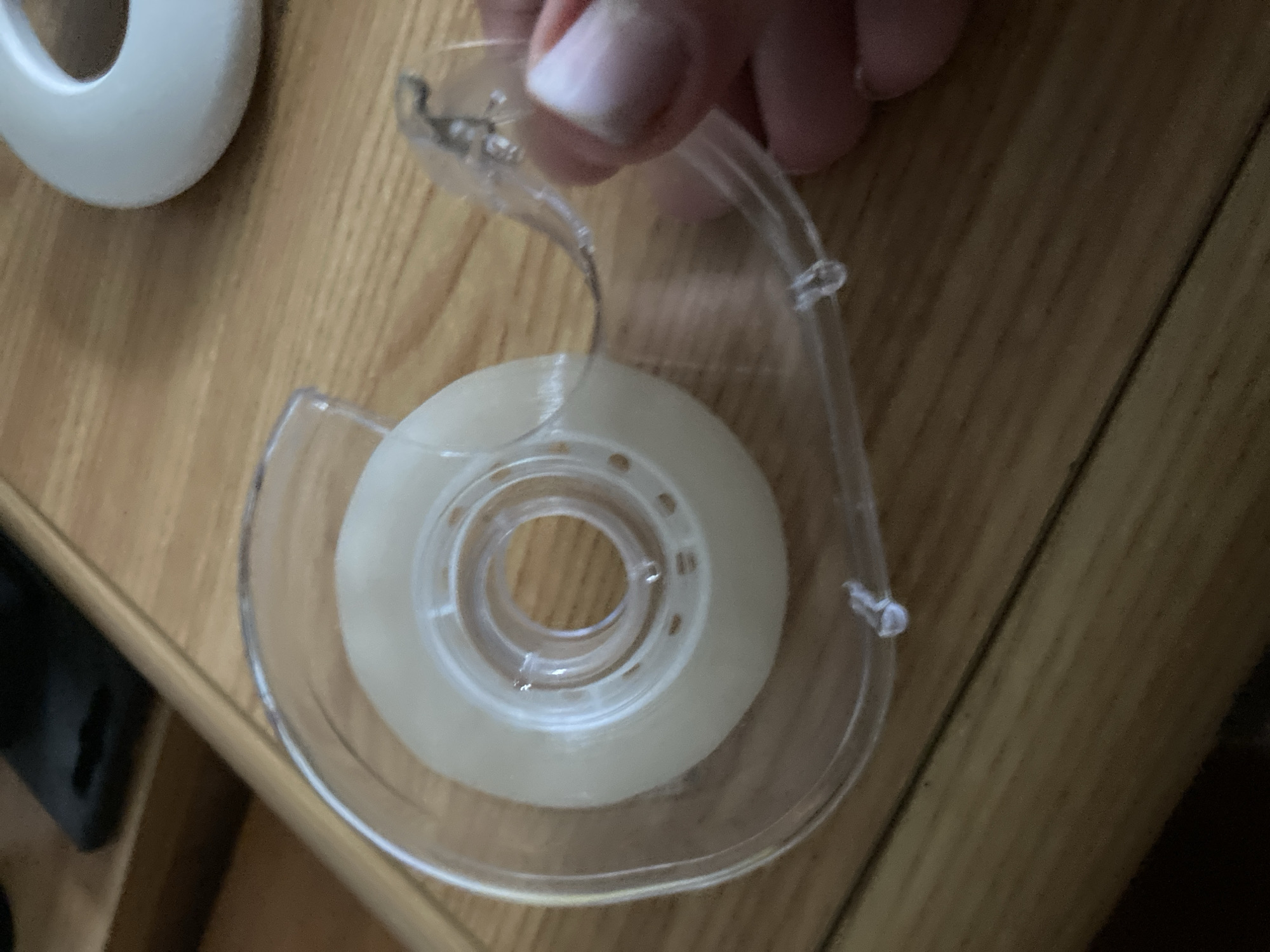 The image shows a close-up of a roll of clear adhesive tape on a wooden surface. A person's hand is visible on the right side of the image, holding the tape dispenser. The tape dispenser is clear plastic and appears to be partially used. The wooden surface has a warm, natural finish and is likely a table or desk. There are no other discernible objects or elements in the image that would suggest a specific location within the city of Lingen (Ems), Germany.
