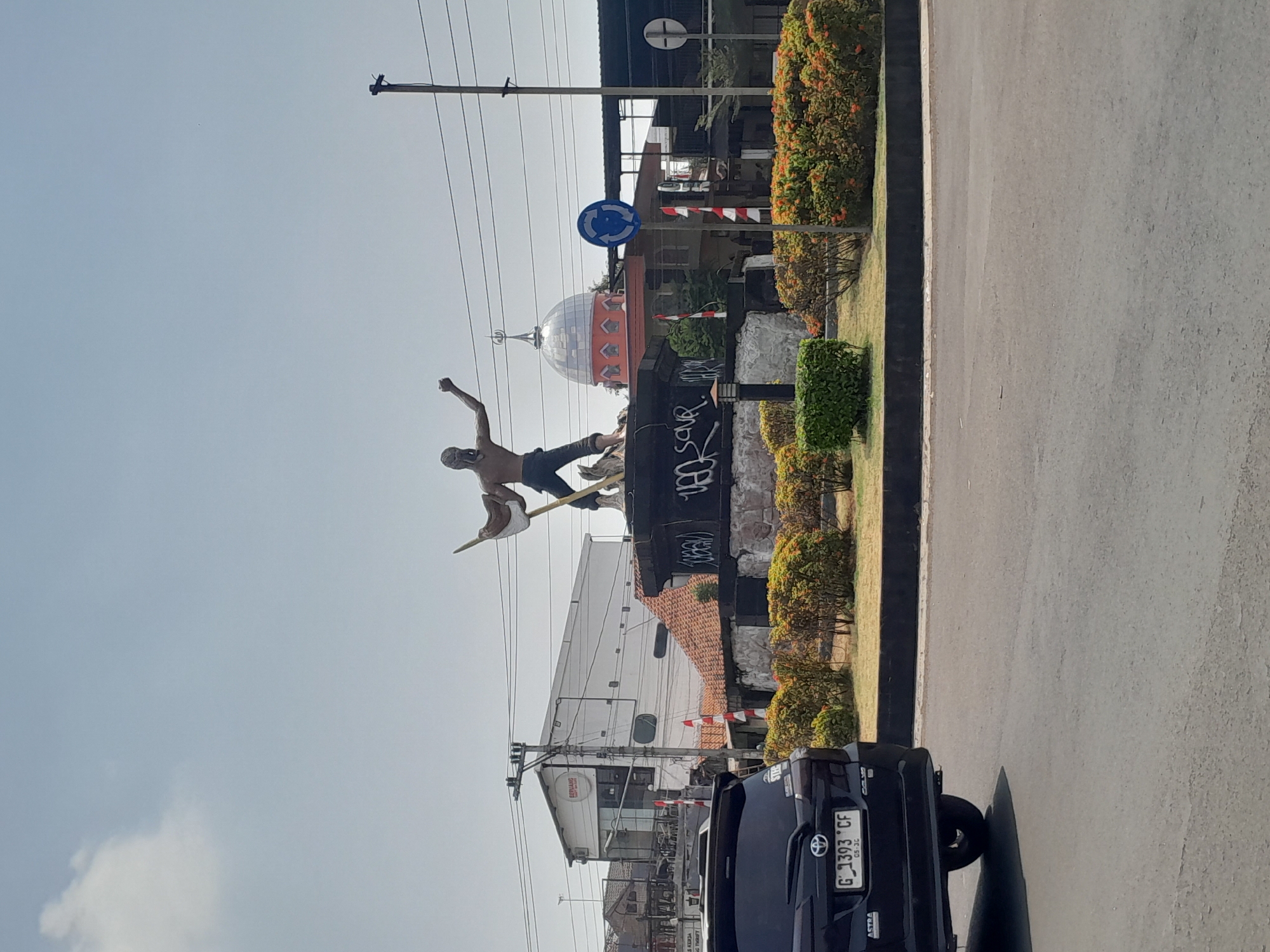 This image captures a street scene in Tegal, Indonesia, centered on a roundabout with a prominent statue. In the foreground, the rear of a dark-colored SUV, license plate G 1393 CF, is visible, facing left and partially obscuring the roundabout's landscaping. The roundabout itself features a circular base for the statue, adorned with graffiti. The statue depicts a male figure, seemingly in motion, holding a spear-like object. Behind the roundabout, a building with a domed roof, likely a mosque, stands with its orange-hued lower section and silver dome contrasting against the sky. Several red and white flags are displayed on poles around the roundabout. Electrical poles and power lines are a recurring element in the background, indicating urban infrastructure. The sky is clear with scattered clouds. The overall environment suggests a public space in a city, likely a traffic intersection.