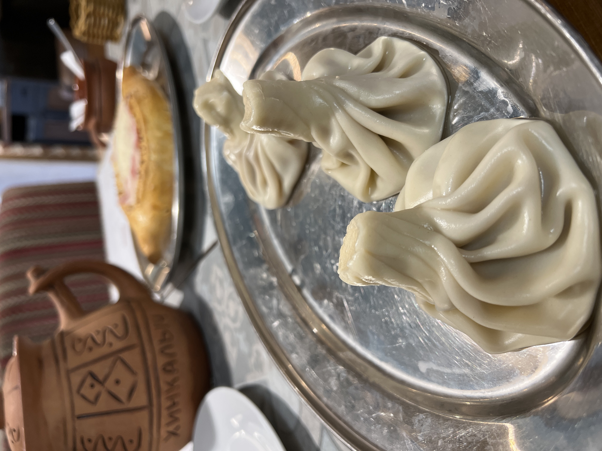 The image presents a close-up, high-angle view of a table in what appears to be a restaurant or cafe in Kazan, Russia. The main subjects are various dishes, typical of Georgian cuisine.
Dominating the right side of the frame is a round, shiny silver metal tray holding several large Khinkali, which are pleated Georgian dumplings. Their dough is light-colored and appears soft and moist, with some liquid visible on the tray around them.
To the left, on a separate, elongated silver metal tray, lies a golden-brown baked good, likely a type of Khachapuri (Georgian cheese bread). It has a long, boat-like shape with a visible light-colored filling, possibly cheese, in its open center.
In the lower-left foreground, a brown, unglazed earthenware pitcher is visible. It features decorative geometric carvings and Cyrillic text along its side. The text clearly reads "ХИНКАЛЬНЫ" (Khinkalny), meaning "Khinkali house" or "Khinkali restaurant," which serves as a strong contextual clue about the dining establishment.
The background is softly blurred, showing glimpses of a striped fabric, possibly a placemat or seat upholstery, and other indistinct table elements. The lighting suggests an indoor setting, indicating a meal is either about to be served or has just been placed on the table. No people are visible in the scene.