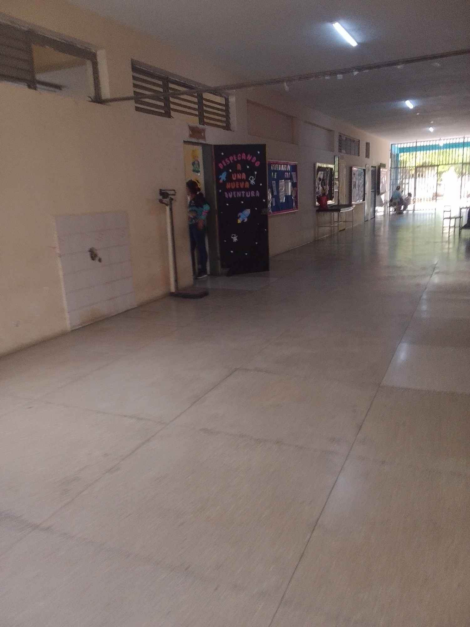 This media file depicts a long, brightly lit hallway in what appears to be a school or community center. The hallway has a light-colored tiled floor and cream-colored walls. Fluorescent lights are visible on the ceiling, illuminating the space.
On the left side of the hallway, there's a cream-colored wall with a white panel featuring some markings, possibly a bulletin board or whiteboard. Next to it, a person, likely a young woman or girl, is standing next to a tall, metallic object, which seems to be a scale. She is wearing a colorful shirt and dark pants. Behind her, on the wall, is a dark rectangular display board with colorful lettering and images, including a rocket and stars. The text on this display reads "DESPEGANDO A UNA NUEVA AVENTURA".
Further down the hallway, several other display boards or bulletin boards are mounted on the walls, some with framed pictures and articles. There's also a table with items on it.
The hallway opens up to an area with a metal gate or bars, allowing natural light to enter. Through the gate, greenery and sunlight are visible, suggesting it is daytime and the weather is pleasant. In the distance, through the gate, a few people are seated, possibly in an outdoor area or waiting room. Two chairs are also visible.
The scene appears calm and orderly, with no indication of any specific event or rushed activity. The overall impression is of a typical day in an educational or community facility in La Tagua, Venezuela. The lighting and the presence of educational displays suggest a daytime setting, possibly during school hours or a community program.