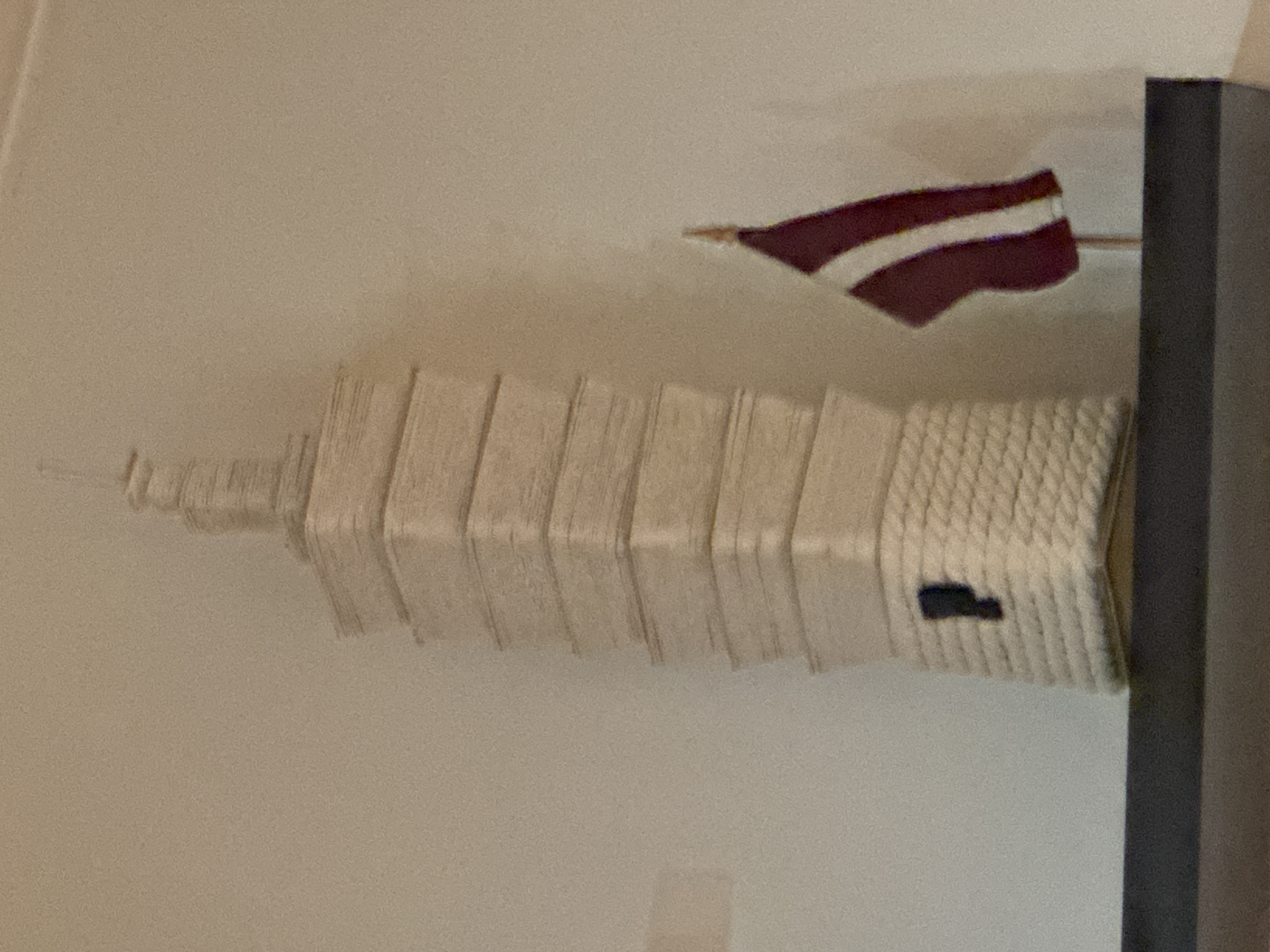 The image shows a vertical stack of multiple stacked structures, resembling a tower, possibly a miniature model of a building. On top of it is a small flag with maroon and white stripes. To the right, there is a dark gray rectangular structure. The background is a light beige wall.
Based on the structure and flag present in the photo, the stack structures could be the tower "Campanario de la Iglesia de San Francisco", located in Puebla, Mexico. The flag resembles the flag of Latvia. Further analysis will be required for complete verification.