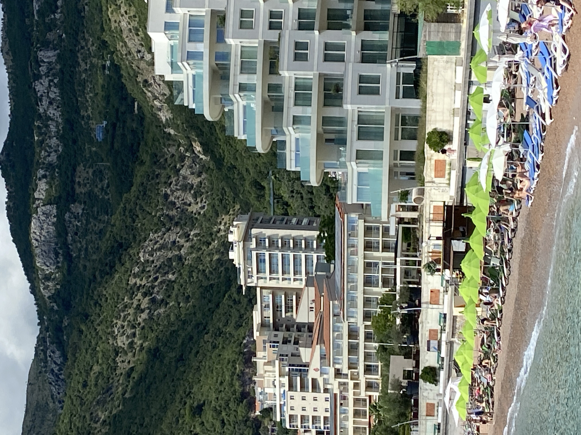 This is a vertical, medium shot image of a beach in Petrovac centar, Montenegro, on a partly cloudy day. The foreground shows a sandy beach with numerous beach chairs and green and white umbrellas. Several people are visible, some lying on chairs and others walking. The water is calm with small waves lapping at the shore.
Behind the beach is a modern, multi-story building with a curved facade, featuring balconies with glass railings. Further inland, a cluster of other apartment buildings of varying heights and architectural styles are visible. The background is dominated by a steep, tree-covered mountainside, with rocky outcrops and a few scattered structures visible on the slopes. A cable car can be seen ascending the mountain in the distance. The scene depicts a popular tourist destination with a blend of natural beauty and modern development.