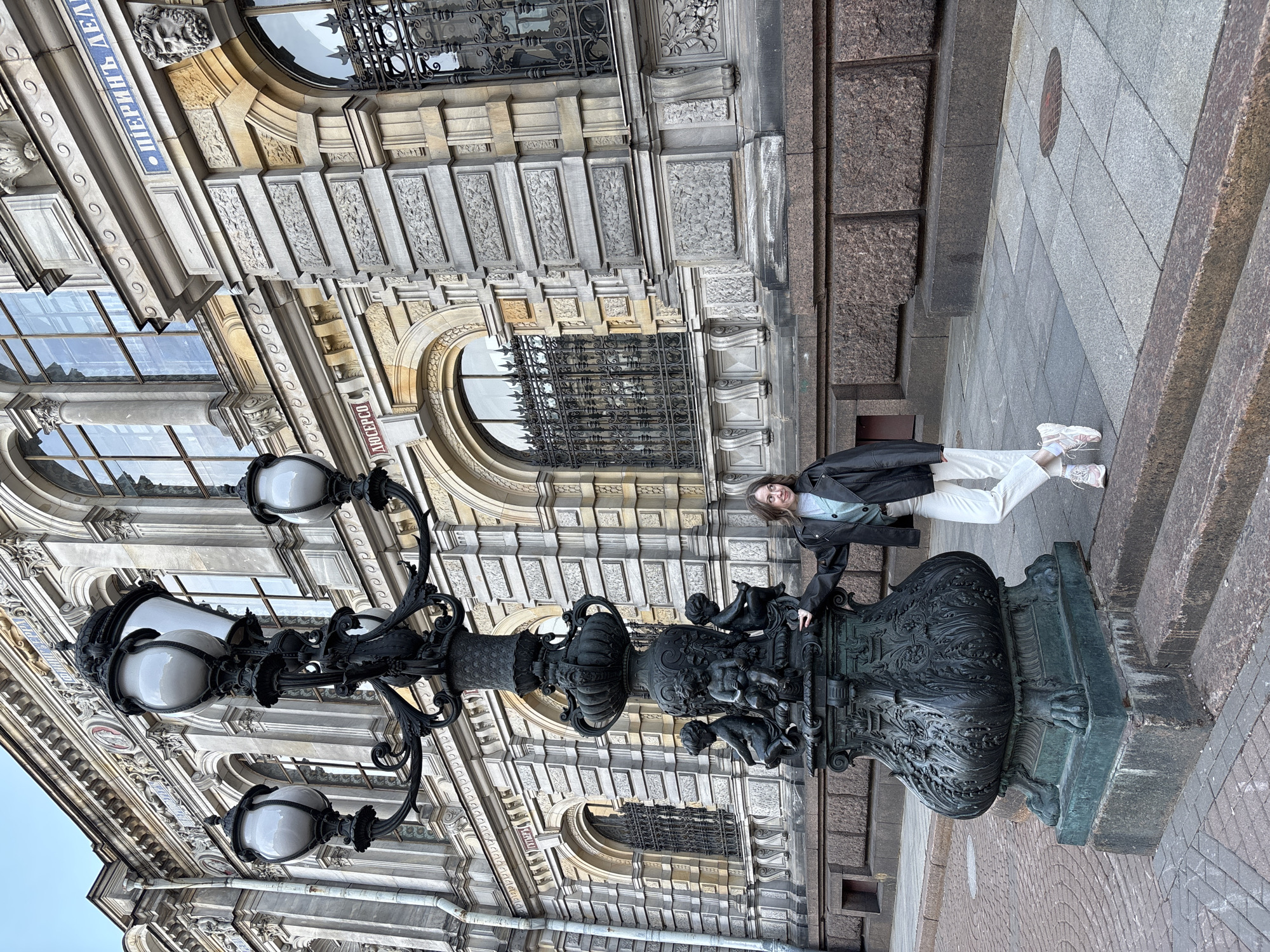 The photograph depicts a young woman standing next to an ornate, cast-iron street lamp in St. Petersburg, Russia. The woman is wearing white pants, a light-colored sweater, and a black leather jacket, and is posing next to the base of the lamp. The lamp has a large, intricately decorated base with cherubic figures, and multiple white globe lights extending from its top. The architectural backdrop features an elaborate facade with arched windows, ornate detailing, and signage in Cyrillic script. The ground is paved with gray and brown stones.