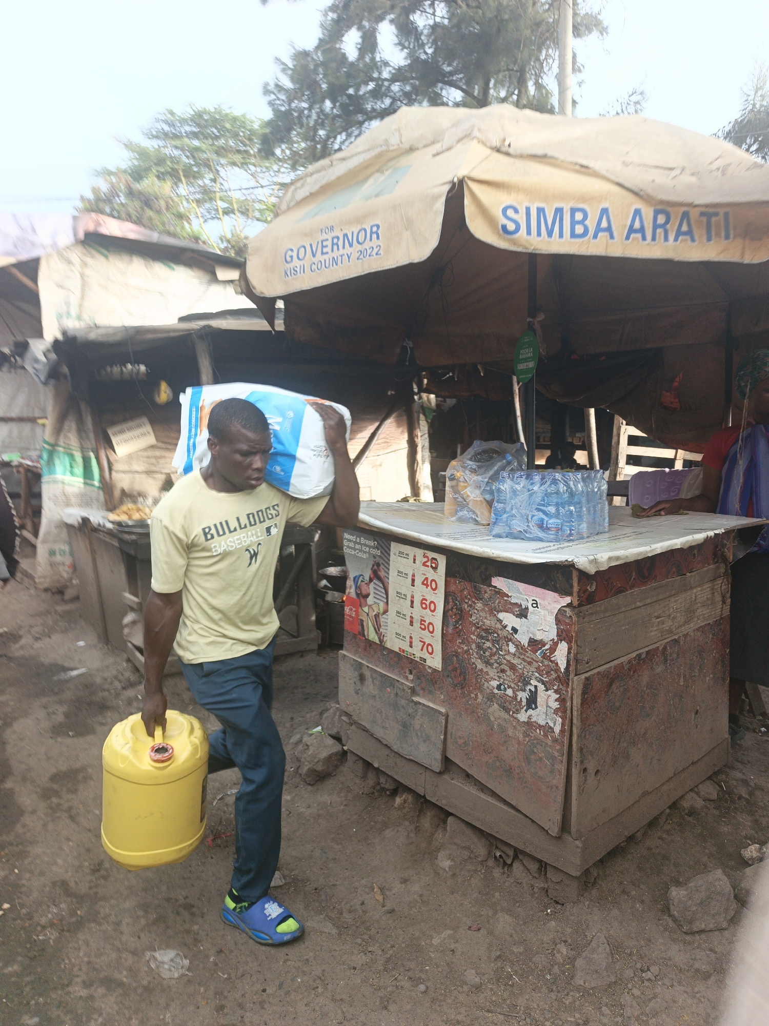 The image depicts a man carrying a large white bag on his left shoulder and a yellow jerrycan in his right hand. He wears a light yellow t-shirt with "BULLDOGS BASEBALL" printed on it, blue trousers, and blue and green sandals.
He is walking past a stall selling bottled water and other goods. The stall is covered by a canopy with the words "SIMBA ARATI FOR GOVERNOR KISII COUNTY 2022" printed on it. A woman in red is present at the store. A price list with prices of drinks is attached to the stall. The stall appears to be constructed from wood.
The ground is dirt, and there are shacks and other buildings in the background. The surrounding environment suggests a low-income or informal settlement in Nairobi, Kenya.