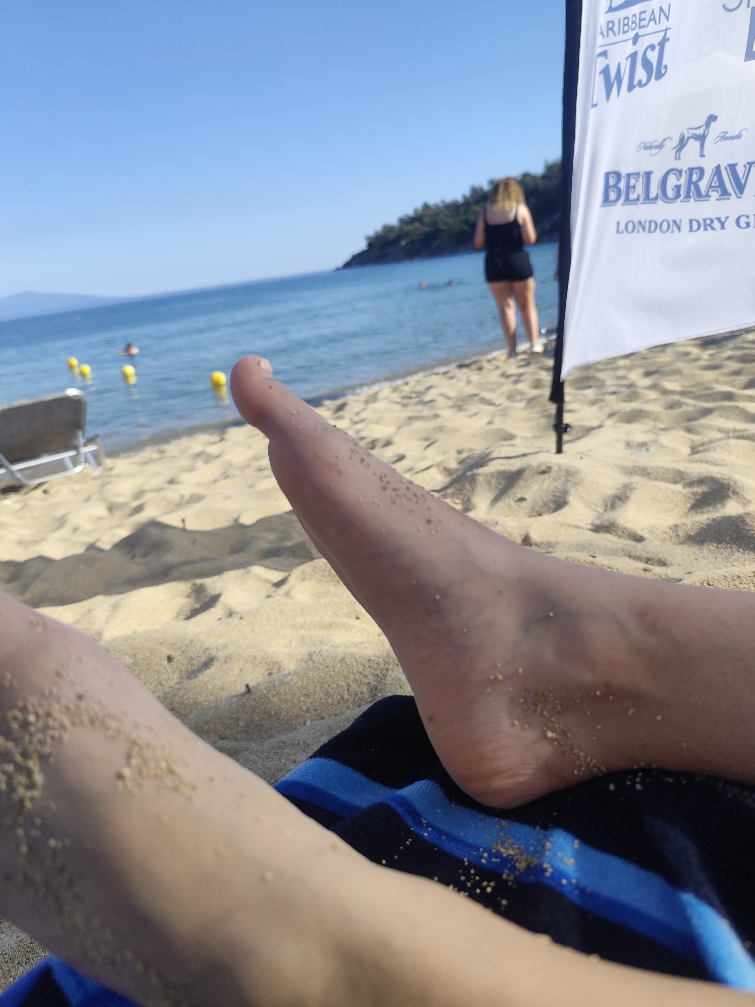 The image shows a close-up view of a person's bare feet on a sandy beach. The feet are covered in fine grains of sand. In the background, the turquoise waters of the Aegean Sea are visible, with a few yellow buoys floating on the surface. A person in a black dress is standing near the water's edge, with their back to the camera. To the right of the frame, a white banner with blue text advertising "BELGRAVIA LONDON DRY GIN" is partially visible, supported by a black pole. The scene is set on a sunny day, with a clear blue sky overhead. The overall atmosphere is one of relaxation and leisure at a beach in Kavala, Greece.