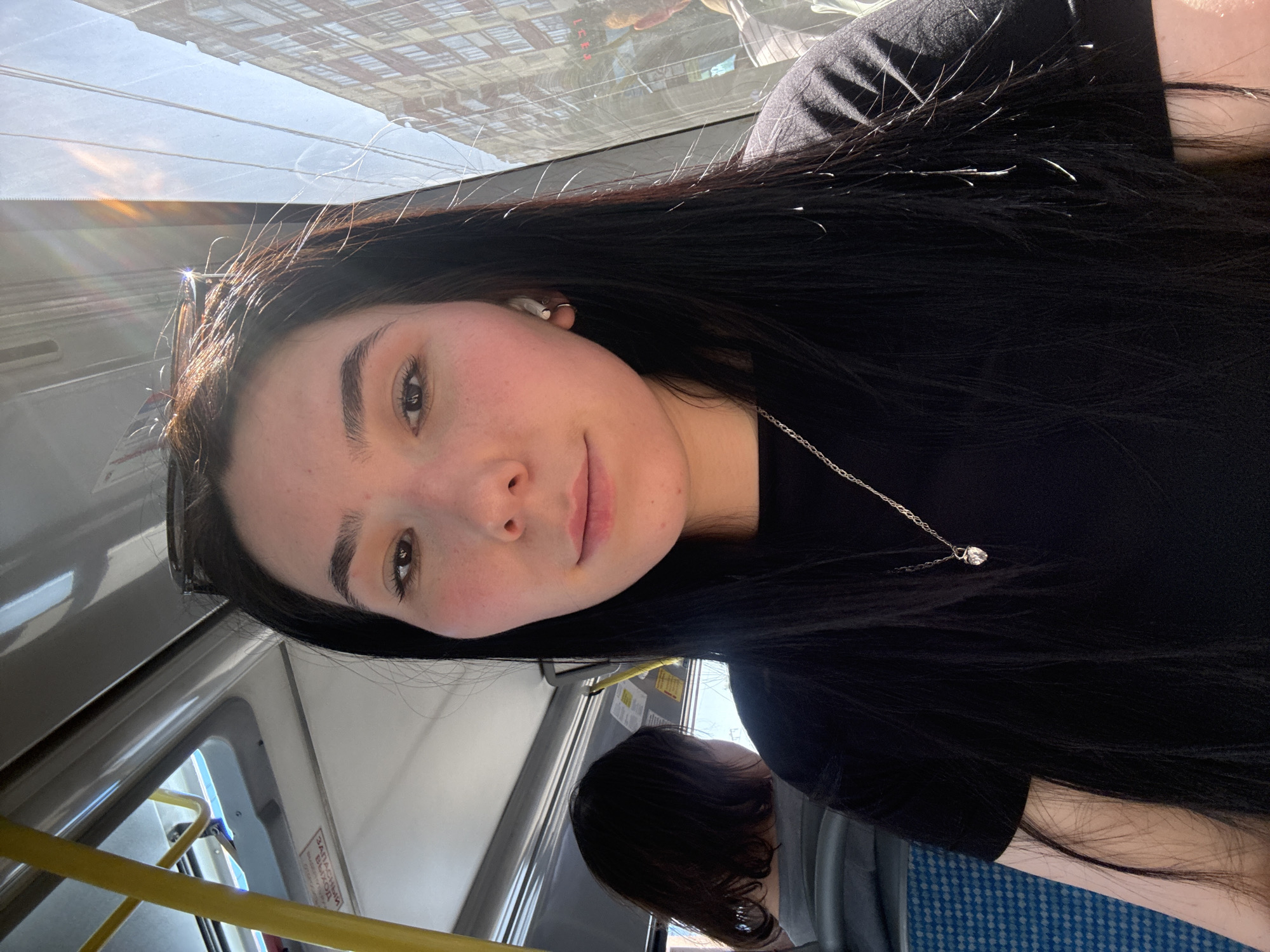 A young woman is seated in a bus. She has dark hair, fair skin, and is wearing a black shirt with a silver necklace. An earbud is visible in her right ear. Behind her, another person with short dark hair is seated. The bus interior features blue patterned seating and yellow handrails. Outside the bus window, the reflection of buildings can be seen, indicating an urban environment. The location is Izhevsk, Russia.