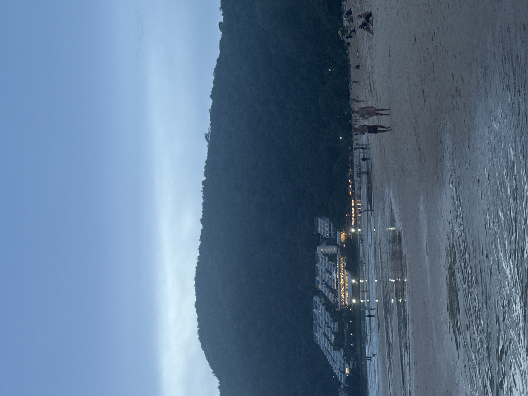 The image depicts a twilight scene on a beach with a gentle tide. In the foreground, the wet sand reflects the dim light, with ripples and patterns from the receding water. Further back, the beach is populated by several individuals walking and sitting, suggesting a relaxed atmosphere. In the middle ground, the ocean meets the shore with small waves. In the background, a hillside covered in lush, dark vegetation rises steeply, with a complex of white buildings nestled on its lower slopes. The buildings are illuminated, their lights twinkling against the darkening landscape. The sky is overcast, with a soft blue-gray hue. The overall mood is peaceful and serene.