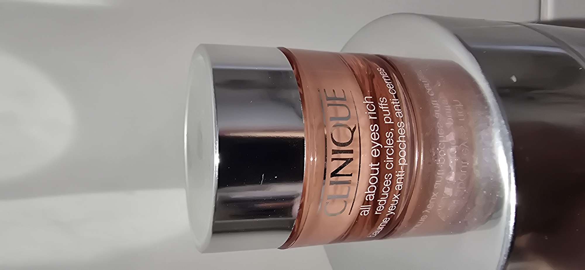A close-up, slightly blurry, eye-level shot shows a Clinique "All About Eyes Rich" cream jar. The jar has a metallic silver lid and a translucent pink base. The label on the jar is white and features the Clinique logo in a metallic font, followed by "all about eyes rich," then "reduces circles, puffs," and finally "baume yeux anti-poches anti-cernes." The jar is positioned on a reflective silver surface, possibly a pedestal or another product container, against a plain white background with subtle shadows. The lighting is even, highlighting the product's packaging.