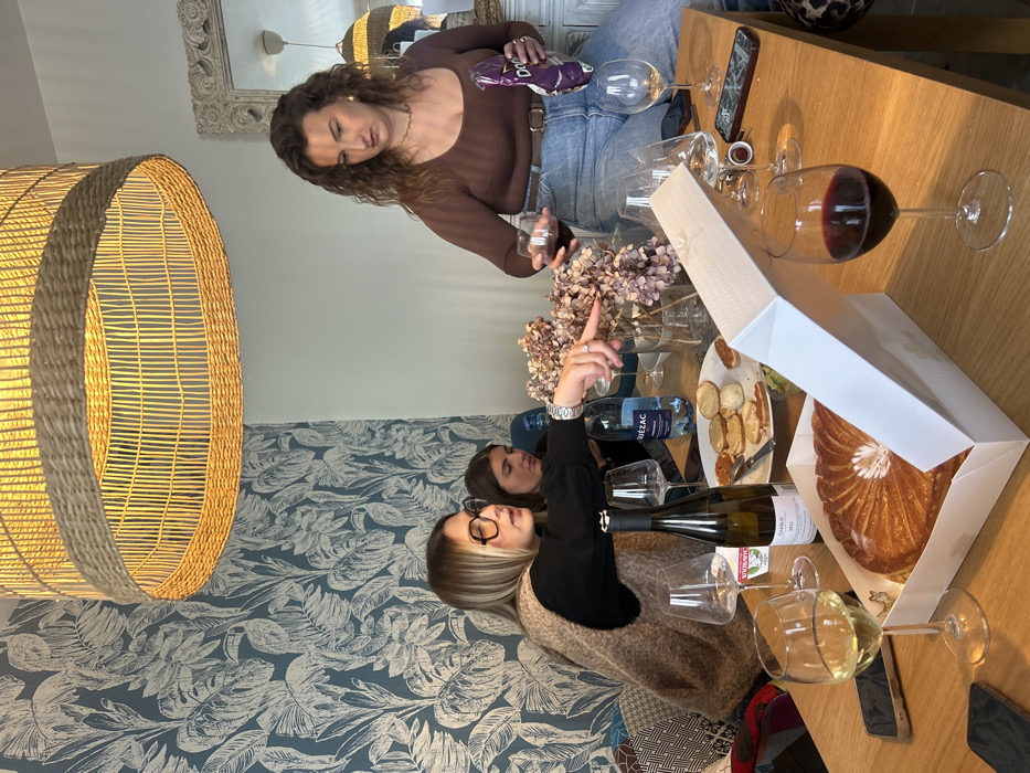 This image depicts an indoor social gathering, likely in a residential dining or living area. While the specific location within Saint-Étienne, France, cannot be determined from visual information alone, the setting is a modern, decorated room.
Three women are present. On the left, a woman with blonde hair and black glasses, wearing a black long-sleeved top and a brown sleeveless vest, is seated at a wooden table. She points with her right hand towards the center of the table, wearing a watch and a ring. Behind her, partially obscured, is a second woman with dark hair, also seated at the table. On the right, a third woman with curly dark hair, wearing a brown long-sleeved top and blue jeans, sits on what appears to be a white cabinet or counter. She holds a purple Doritos bag in her left hand and a wine glass in her right, seemingly in the act of pouring or preparing a drink.
The wooden table is central to the scene, laden with various items. Prominently displayed is an open white cardboard box containing a Galette des Rois (King's Cake). Nearby are multiple wine glasses, some empty, one containing white wine, and another with red wine. A bottle of white wine, labeled "CHABLIS 2021," stands on the table, alongside a bottle of "QUEZAC" mineral water. A white plate holds an assortment of small appetizers or biscuits, and a small green dish is also visible. Several smartphones are scattered across the table surface. A glass vase containing dried, light-colored hydrangea-like flowers is positioned near the center.
The room features a patterned wallpaper on the left wall with blue and white tropical leaf motifs. Overhead, a large, cylindrical pendant light fixture made of woven rattan casts a warm glow. On the right wall, a white, ornate-framed mirror reflects a portion of the room, including another hanging light fixture. Seating on the left side of the table is adorned with cushions featuring geometric patterns.