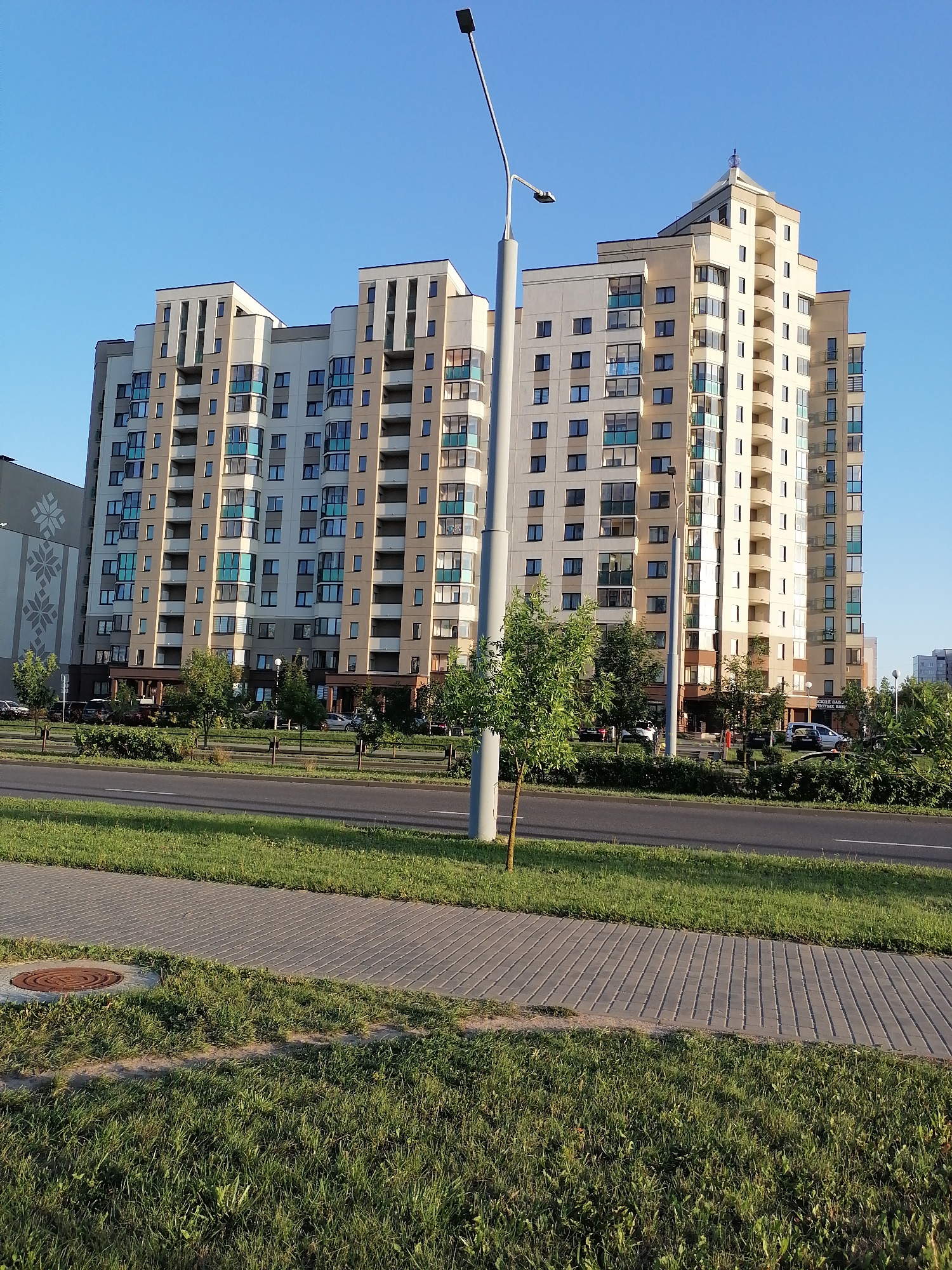 The image depicts a modern, multi-story apartment building in Hrodna, Belarus, under a clear blue sky. The building is light beige with sections of white and light gray accents. It features numerous balconies and windows, some with teal-colored trim. A tall, gray lamppost stands in front of the building, and several trees with green foliage are visible. A paved sidewalk runs along the foreground, bordered by a strip of grass and a road. Cars are parked along the street in front of the building, and a large, patterned structure is partially visible to the left of the apartment building. The scene appears to be a residential area on a sunny day.