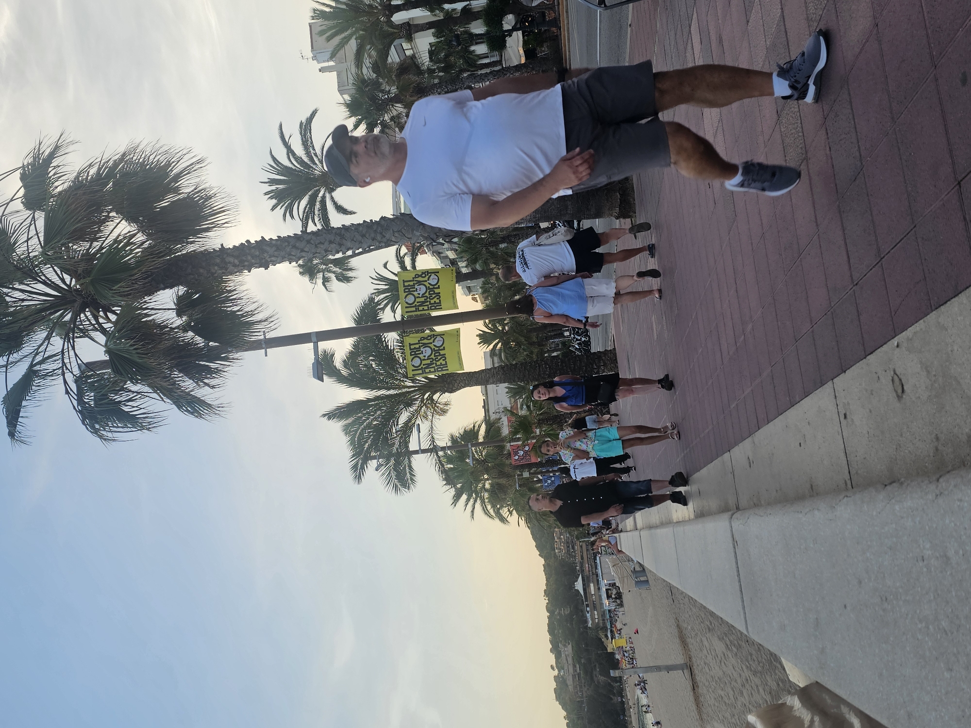The image captures a sunny day scene along a pedestrian walkway bordering a beach in Lloret de Mar, Spain. Palm trees are prominent throughout the frame. In the foreground, a man wearing a white t-shirt and grey shorts is walking towards the right. Further back, several other individuals are also walking, including a woman in a blue top and black skirt, a young girl in a blue top and shorts, a man in a black shirt and shorts, and a couple wearing a white t-shirt and a blue top with white shorts. Banners with text are visible hanging from poles along the walkway, some appearing to be promotional for the town. To the left, a sandy beach is visible with numerous people relaxing and sunbathing, and buildings are situated on a hillside in the distance. The sky is clear with a warm, slightly hazy glow.