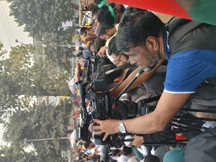 A crowded urban outdoor scene in Dhaka, Bangladesh, centers on a male videographer actively operating a professional video camera mounted on a tripod. The videographer, positioned in the foreground and facing right, is a middle-aged man with dark hair and a short salt-and-pepper beard, wearing a blue polo shirt with white trim on the sleeves and a dark vest. A silver wristwatch is visible on his left wrist as he holds the camera's handle and looks upwards. The camera, a dark-colored professional model, features a prominent lens and a microphone attached via a boom arm extending towards the front.
Surrounding the primary subject and extending into the dense background is a large gathering of predominantly male individuals. Many are standing, some wearing white skullcaps, while others in the middle-ground sport green headbands. Several other people are visible holding cameras or smartphones, suggesting a media presence or participants documenting an event.
The environment depicts a public thoroughfare or square, characterized by numerous trees with varying foliage density, some appearing bare-branched, indicative of an urban park or tree-lined street. Buildings are discernible in the distance behind the trees. Faintly visible in the mid-background are market stalls with colorful umbrellas or canopies. A street light pole is also present. A red and green flag, possibly the national flag, is partially visible on the far right edge of the frame. The overall scene suggests a significant public event or gathering requiring extensive media coverage.
