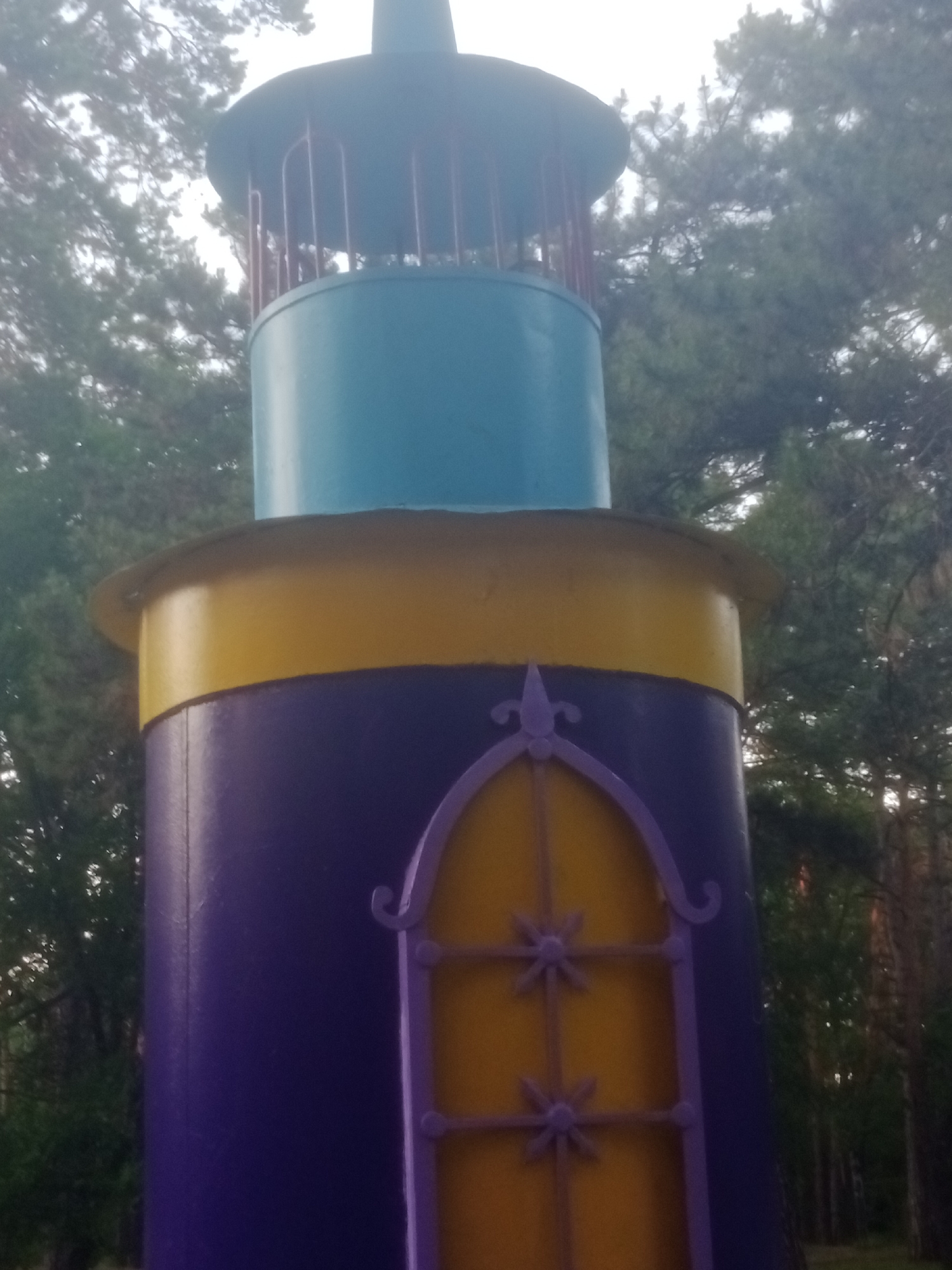 The image shows a close-up of a colorful, decorative tower structure, likely a playground element or art installation, situated outdoors among trees. The tower is painted in sections: the bottom is a deep purple with a decorative yellow window outlined in purple. Above the purple section is a wide yellow band, followed by a light blue cylindrical section. The very top features a darker conical structure with vertical red bars and a dark cap. The background is filled with blurred green foliage of trees, suggesting a park or natural setting. The lighting appears somewhat dim, possibly indicating an overcast day or early morning/late evening. The specific location in Horishni Plavni, Ukraine, is not visually identifiable from the image itself.