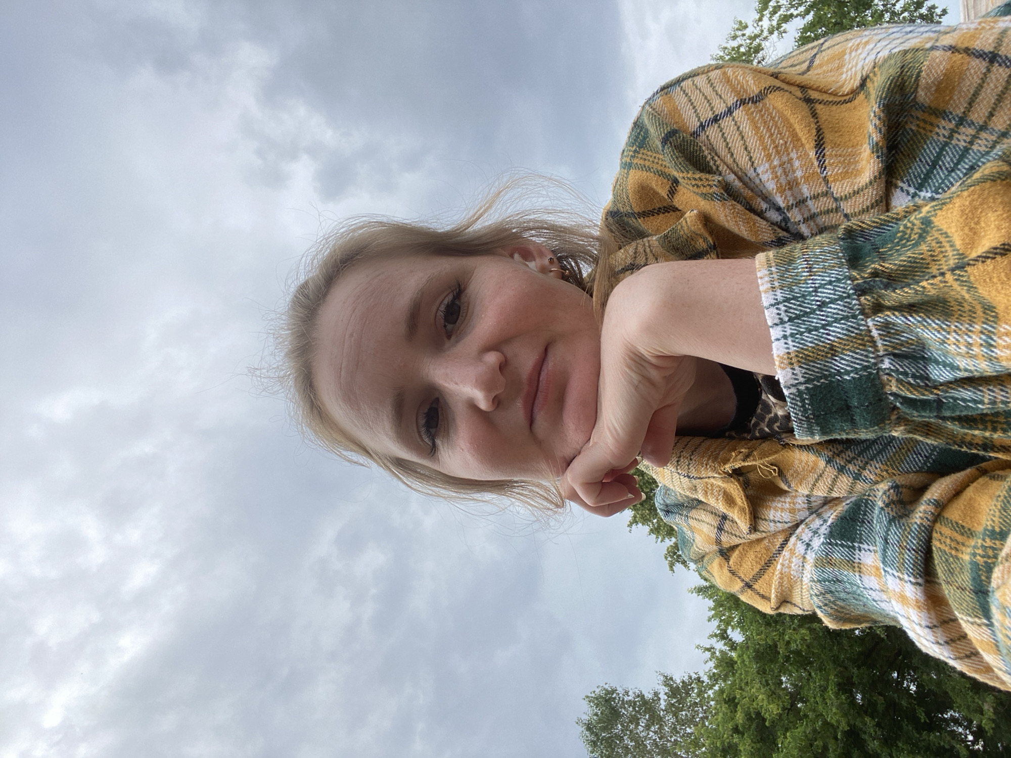 A low-angle, close-up selfie shows a young woman with blonde hair resting her chin on her hand, looking at the camera. She is wearing a yellow, green, and white plaid flannel shirt. Her expression is neutral. The background is an overcast sky with some clouds visible, and the tops of green trees can be seen on the right side of the frame. The lighting suggests it is daytime, likely with cloudy weather. There is no visible text in the image.
