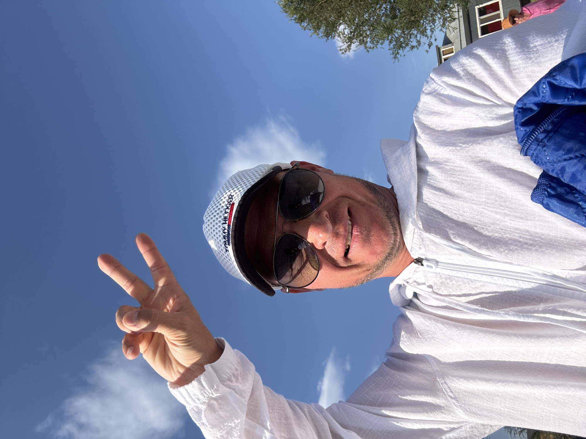 A man wearing a white zip-up hooded jacket, sunglasses, and a white baseball cap is captured in a selfie against a bright blue sky with scattered clouds. His left hand is raised, forming a peace sign with his index and middle fingers extended, while his thumb touches his ring finger and pinky finger. The man has stubble and is smiling broadly. In the background, partially visible, is a tree and some buildings. The location is not identifiable as Urdzhar, Kazakhstan.