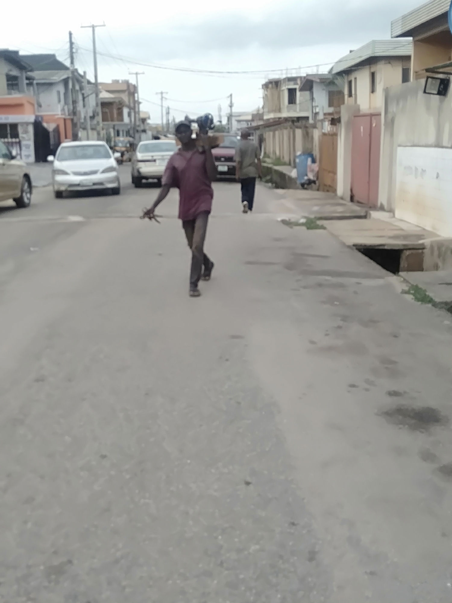 The image shows a street scene in Shangisha, Nigeria. A dark-skinned man walks toward the camera, carrying items over his shoulder. He is wearing a red shirt and dark trousers. Another man walks away from the camera in the same direction. Several cars are parked along the street, and buildings are visible on either side. The sky is overcast.