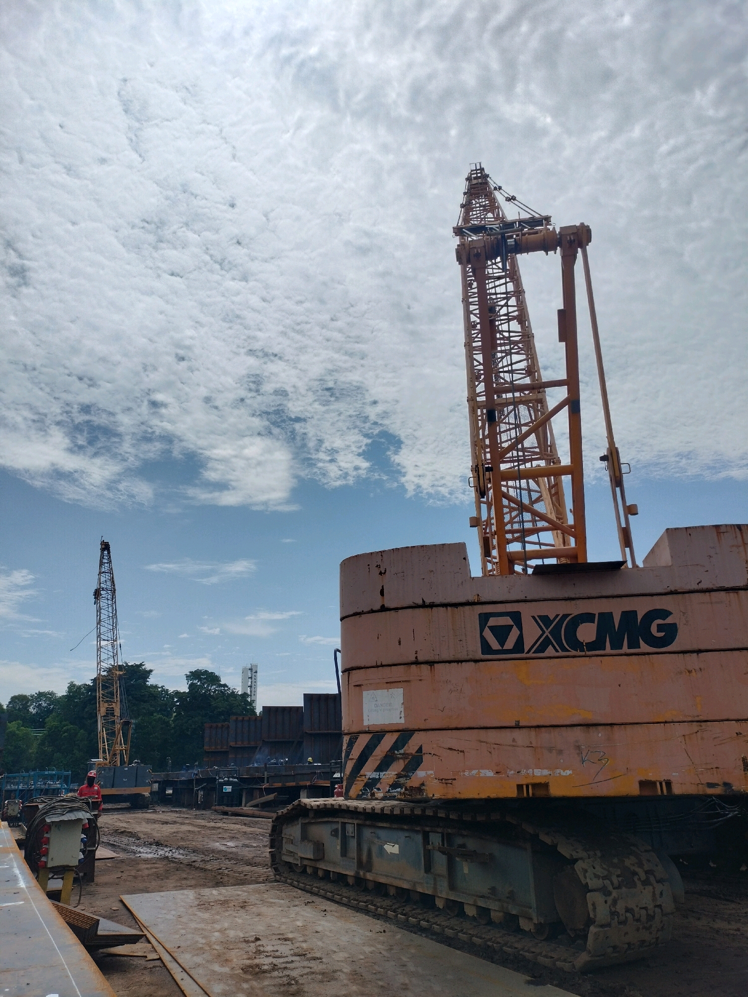 A construction site in Batam, Indonesia, is depicted under a cloudy sky. In the foreground, a large orange and grey crawler crane dominates the right side of the frame, its massive tracks resting on the muddy ground. The side of the crane displays the "XCMG" logo. To the left, another yellow lattice boom crane stands tall, its boom reaching towards the sky. In the middle ground, a person in a red hard hat and vest is visible near some equipment. The area is a muddy, dirt-covered construction zone, with stacks of dark metal containers and other construction materials scattered around. Trees are visible in the background, and a tall white tower structure can be seen in the distance. The overall scene suggests ongoing heavy construction work.